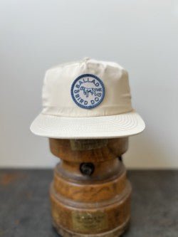 The Shop Hat | Circle Clubhouse Tradition by Ballad of the Bird Dog is a beige baseball cap with a blue patch logo, displayed on a wooden hat stand against a plain background.