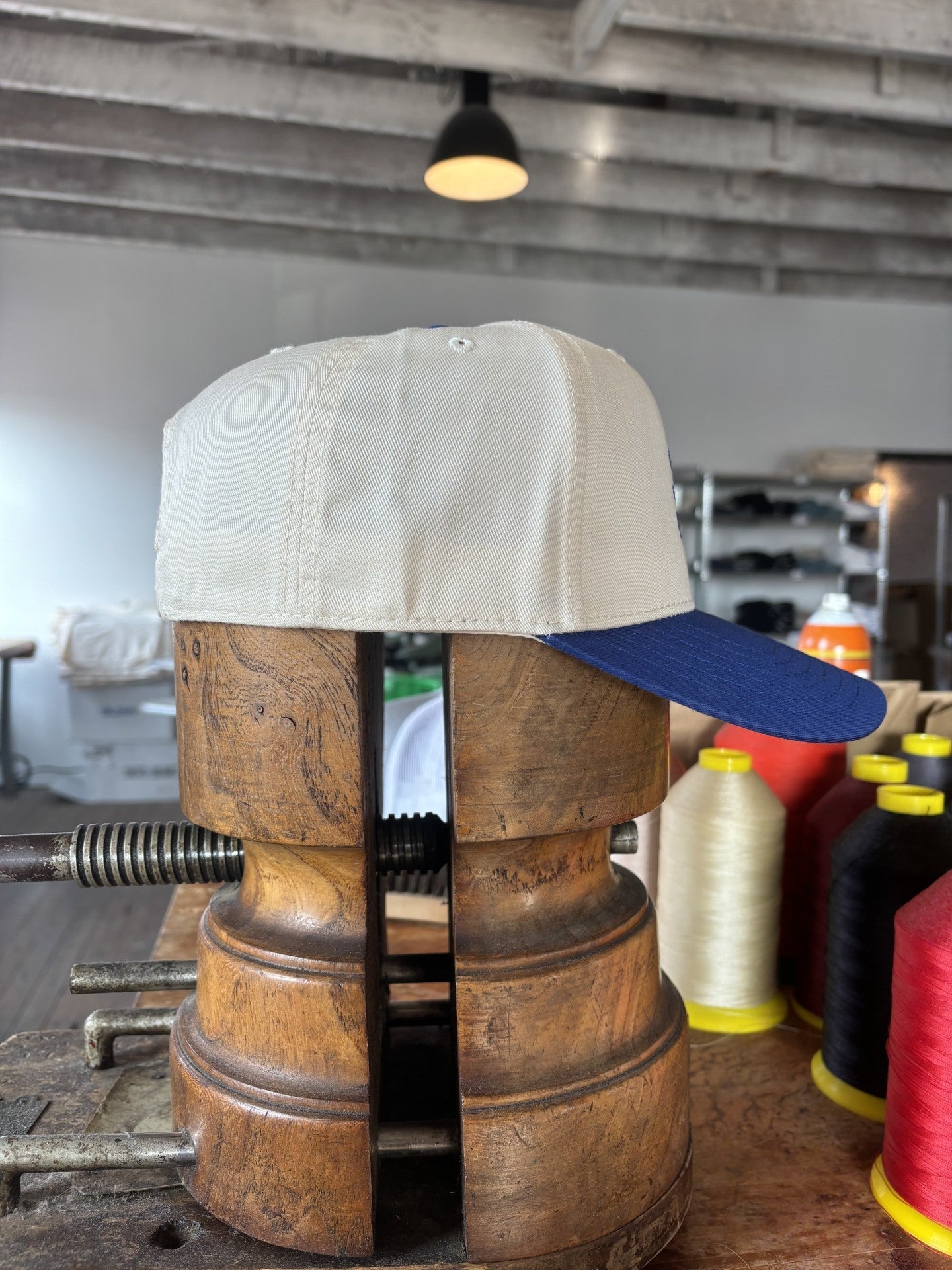 The Shop Hat | Field Companion by Ballad of the Bird Dog—a beige and blue baseball cap—is secured in a wooden hat block clamp, surrounded by spools of thread and shelves in the background.