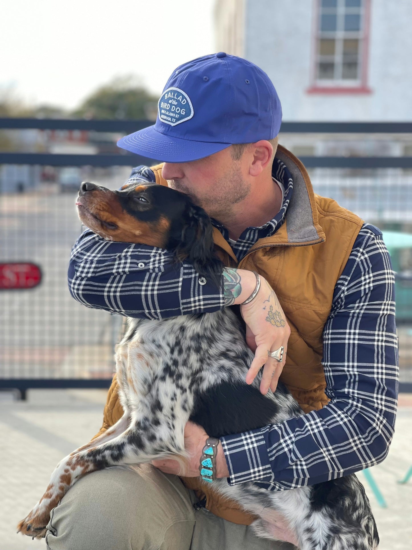 A man wearing the Ballad of the Bird Dog Shop Hat | Home Base hugs and kisses a black and white dog while sitting outdoors in front of a metal fence.