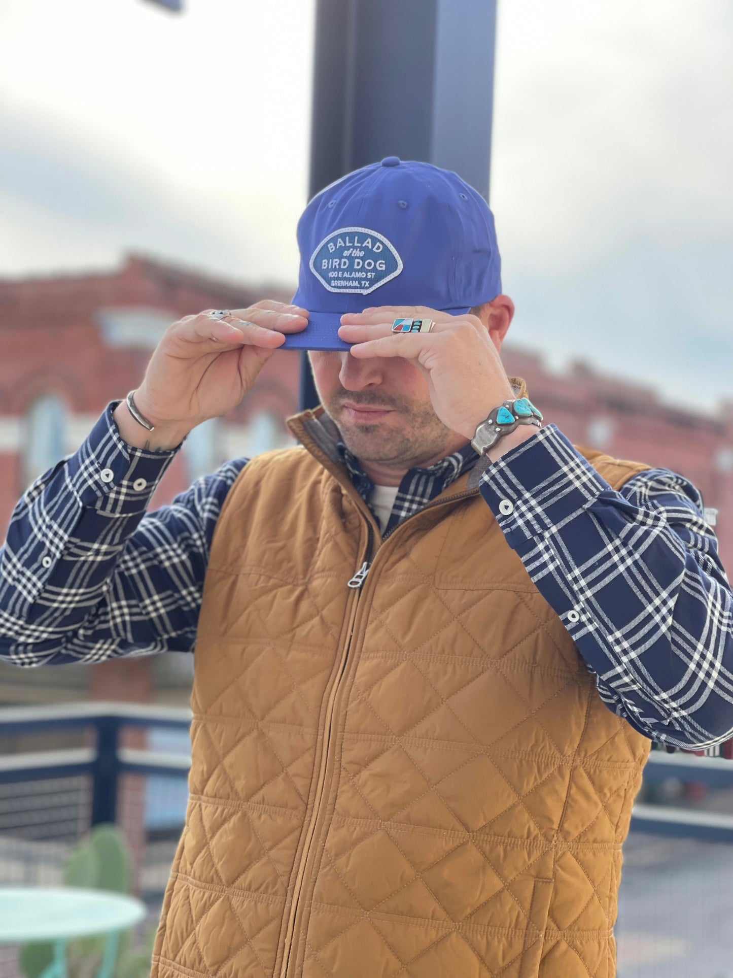 A person outdoors adjusts a blue Shop Hat | Home Base by Ballad of the Bird Dog, paired with a plaid shirt and tan quilted vest. A brick building and railing can be seen in the background.