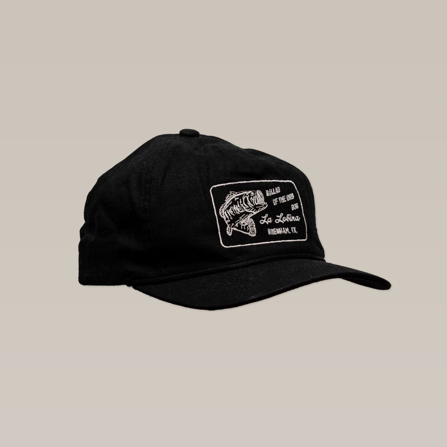 The Ballad of the Bird Dog La Lobina Shop Hat is a black fishing cap featuring a white embroidered fish logo and text patch on the front, shown against a neutral background.
