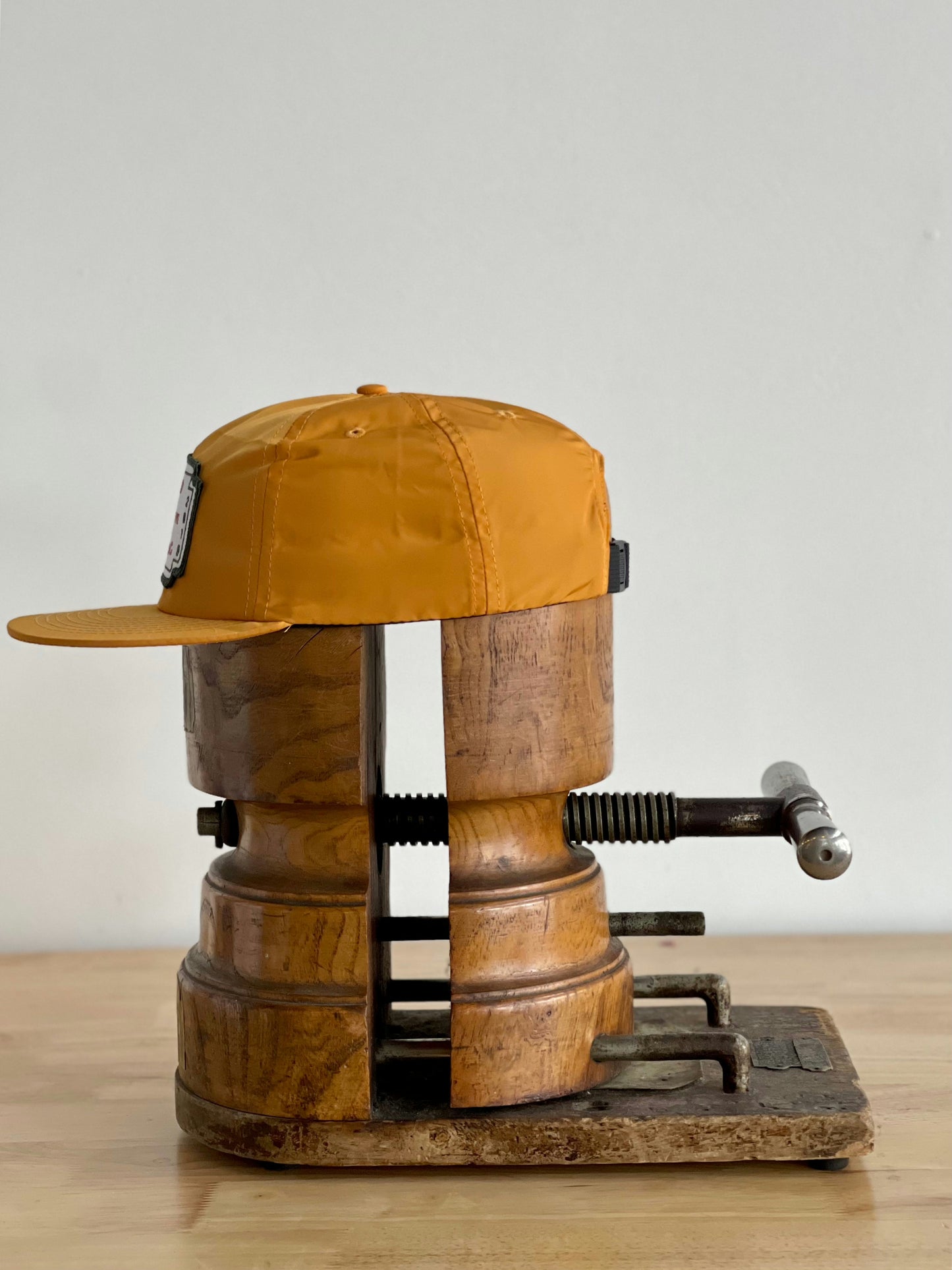 The Shop Hat | Neon Revival by Ballad of the Bird Dog is clamped between wooden blocks in a metal vise on a wooden table, set against a plain white wall—evoking the downtown Brenham vibe.