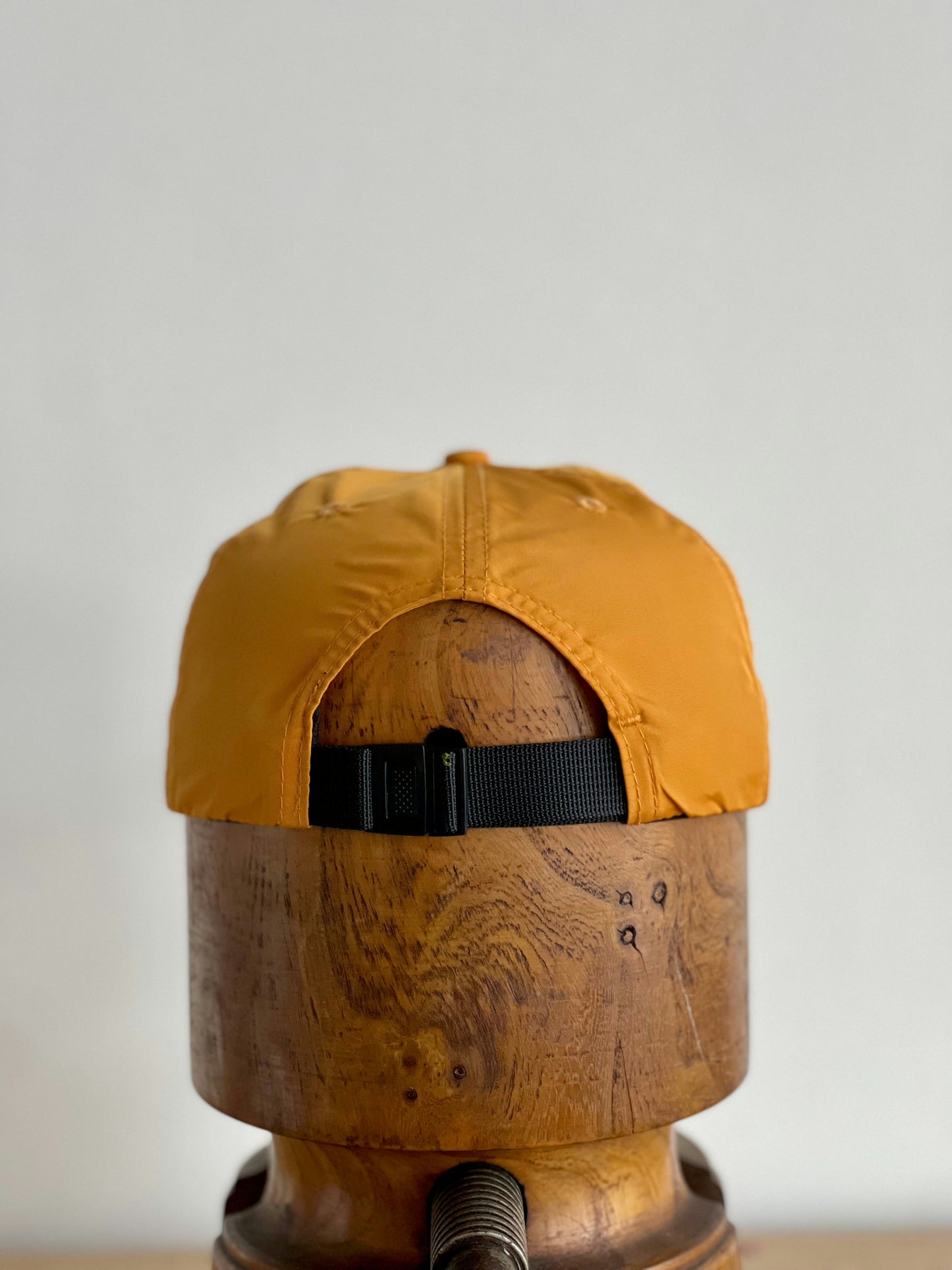 A mustard yellow Shop Hat | Neon Revival by Ballad of the Bird Dog with a black adjustable strap is displayed on a wooden stand from the back—ideal for exploring downtown Brenham in style.