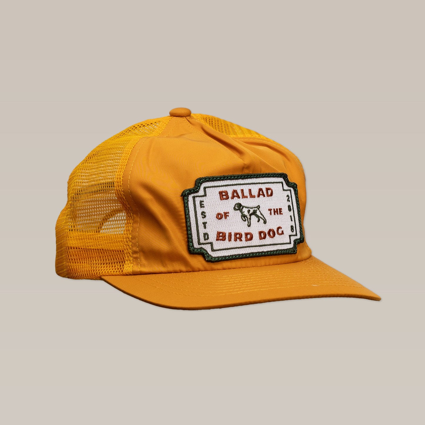The Shop Hat | Neon Revival by Ballad of the Bird Dog is a yellow mesh trucker hat with a front patch reading "Ballad of the Bird Dog" and an illustrated dog, inspired by downtown Brenham, shown on a beige background.