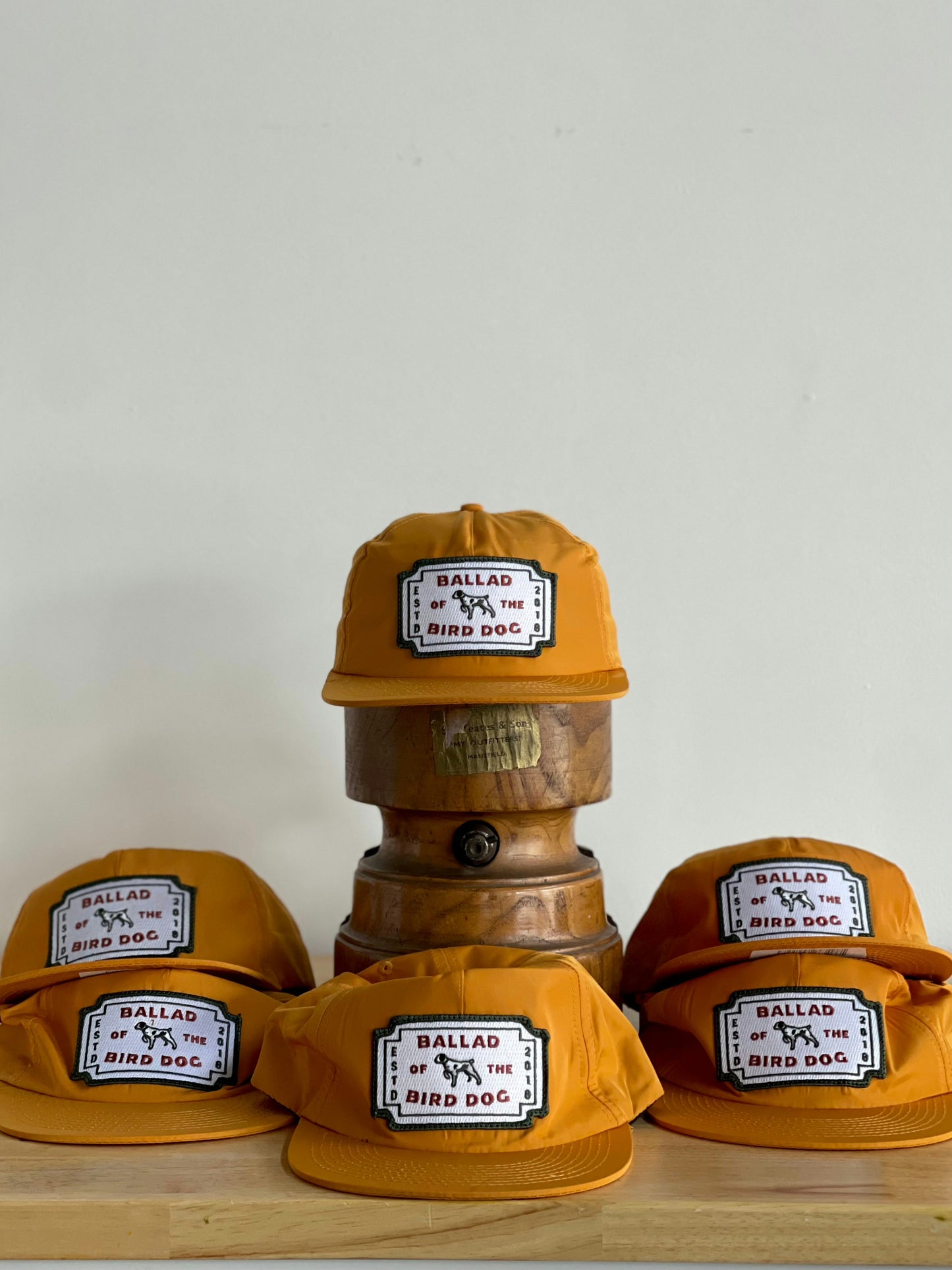 Five Shop Hat | Neon Revival hats by Ballad of the Bird Dog are arranged on a wooden barrel and surface, their bold orange color and signature patches reflecting the lively vibe of downtown Brenham against a plain white background.