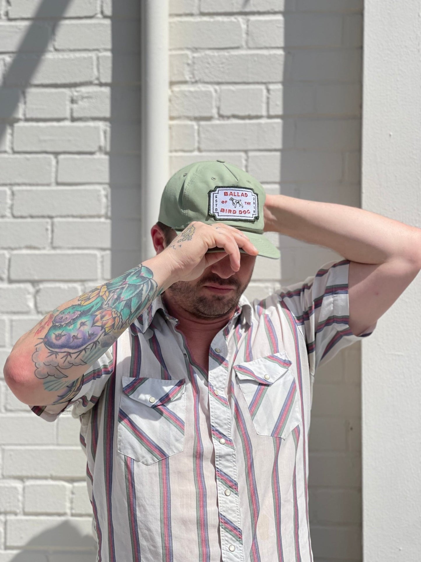 A person with a colorful tattoo adjusts their Ballad of the Bird Dog Shop Hat | Neon Revival, standing against a white brick wall in a striped short-sleeve shirt, reflecting the lively spirit of downtown Brenham.