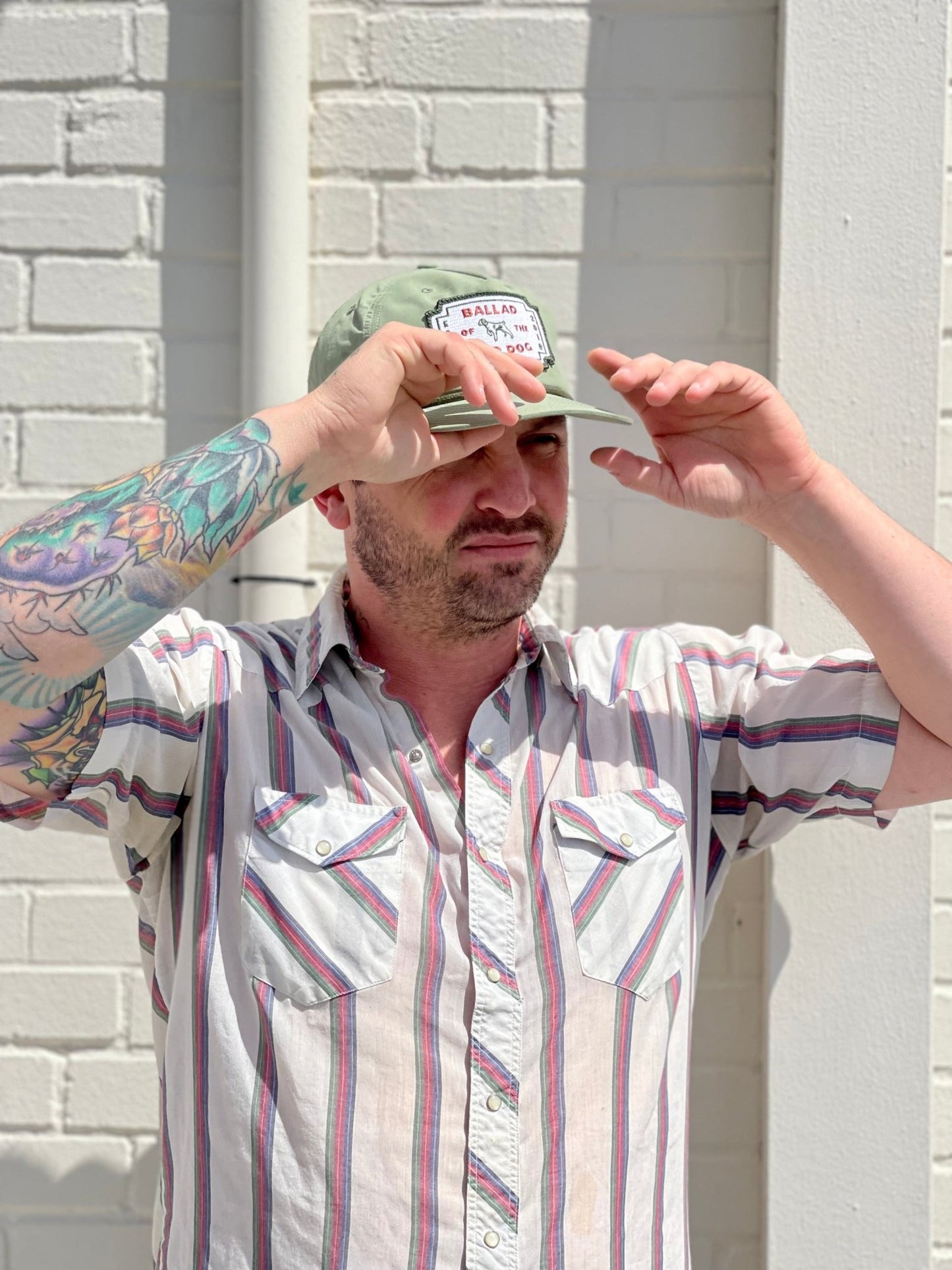 A man with a tattooed arm, dressed in a striped short-sleeve shirt and the Shop Hat | Neon Revival by Ballad of the Bird Dog, stands against a white brick wall, shading his eyes with one hand.