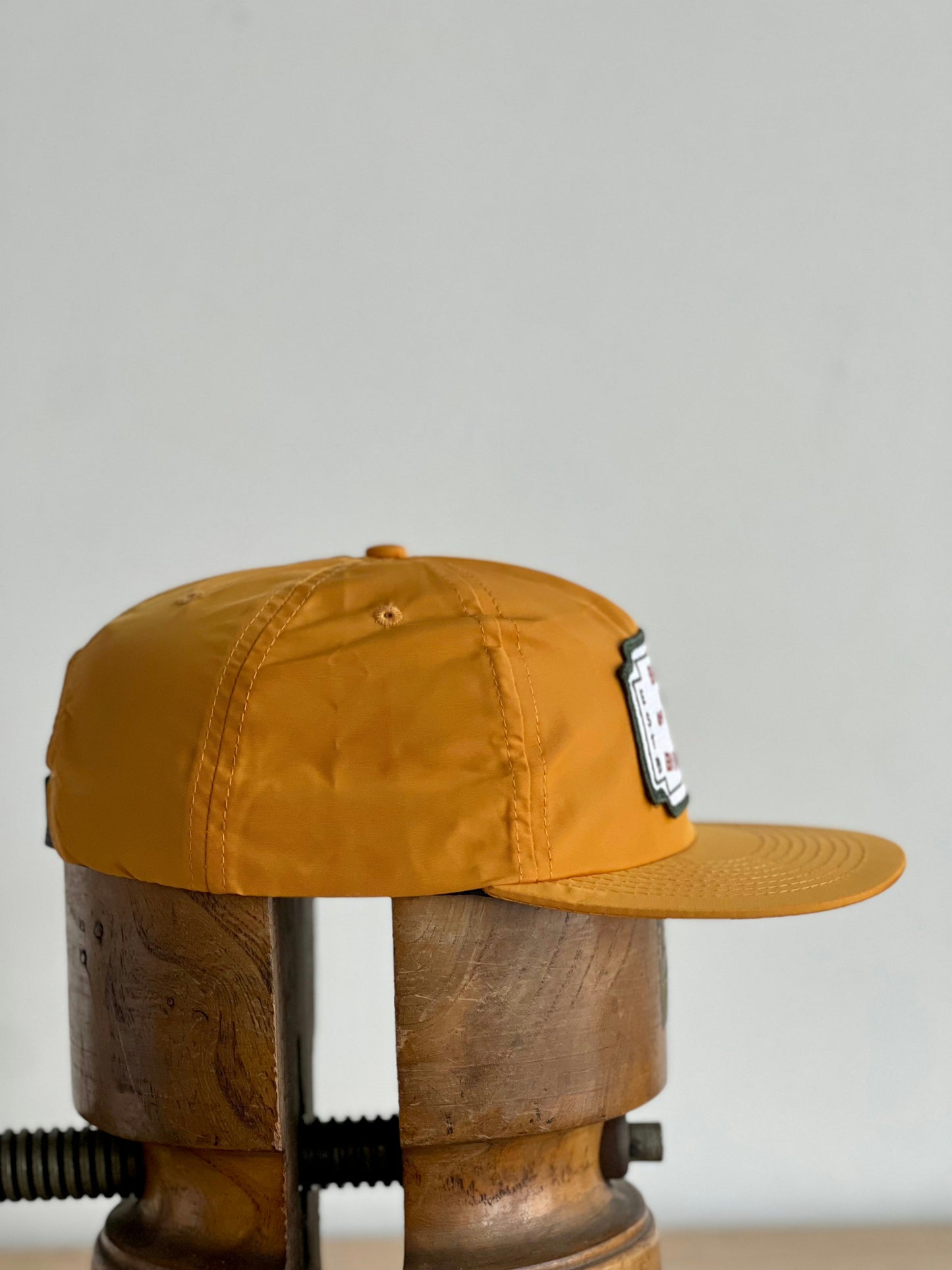 The Shop Hat | Neon Revival by Ballad of the Bird Dog features a classic logo patch in mustard yellow, displayed on a wooden block with a plain light backdrop—an ideal accessory to showcase your style in downtown Brenham.