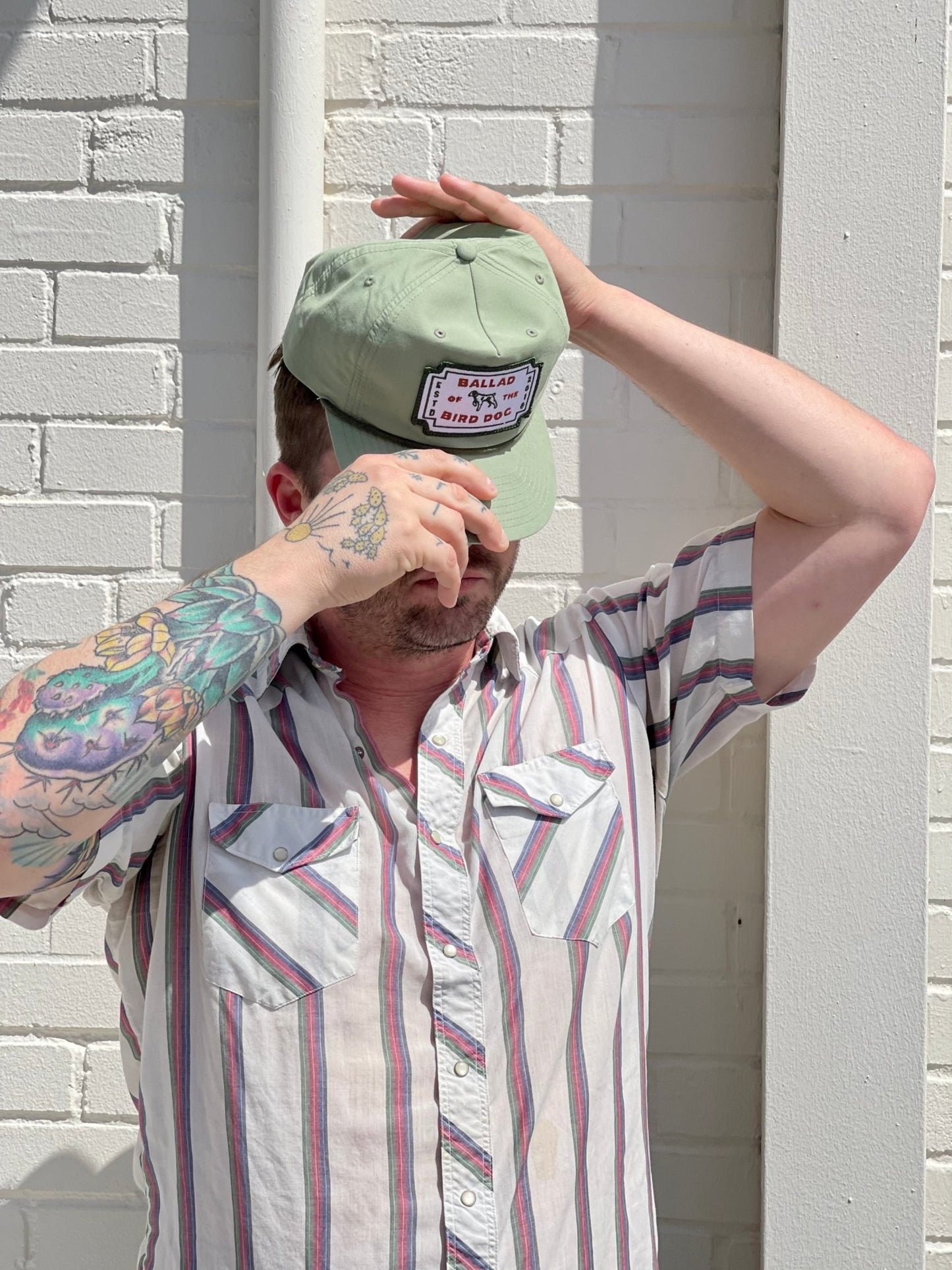Wearing a striped short-sleeve shirt and a Ballad of the Bird Dog Shop Hat | Neon Revival, a person stands before a white brick wall, adjusting the hat with one hand while covering part of their face with the other.