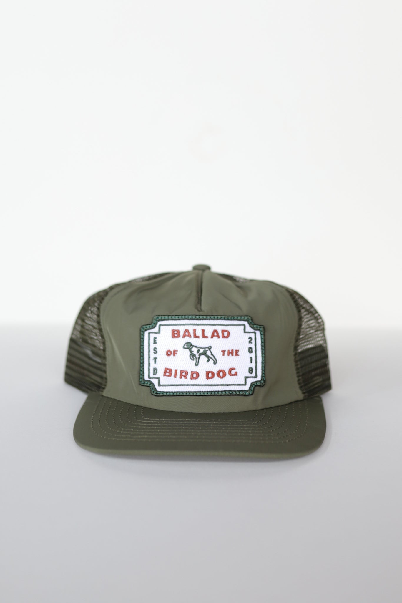 The Shop Hat | Neon Revival by Ballad of the Bird Dog is an olive green mesh-back trucker hat with a front patch featuring a dog and "Ballad of the Bird Dog"—a classic logo inspired by downtown Brenham.