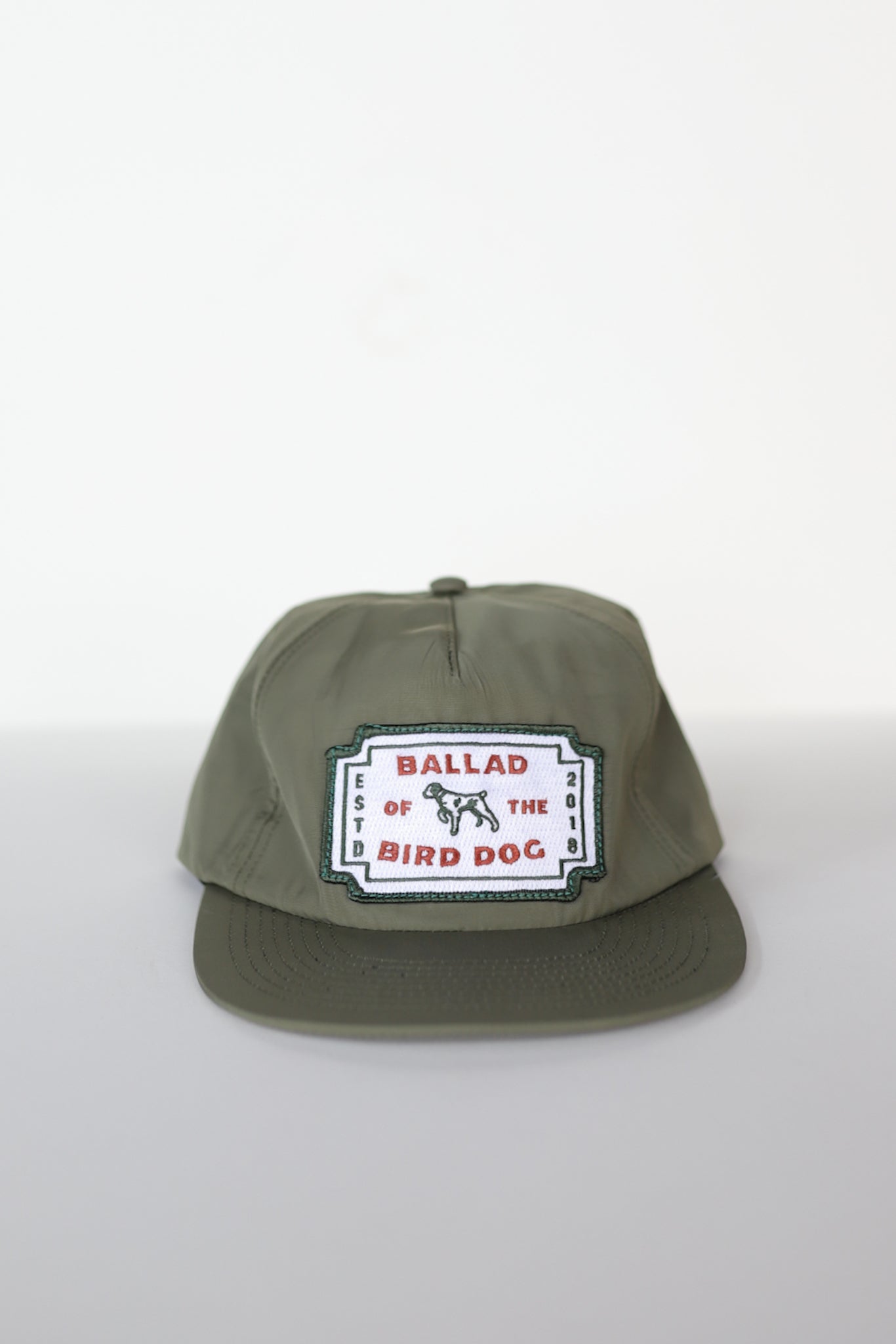 The Ballad of the Bird Dog Shop Hat | Neon Revival features a classic olive green design with a rectangular logo patch and dog illustration—ideal for your next stroll through downtown Brenham.