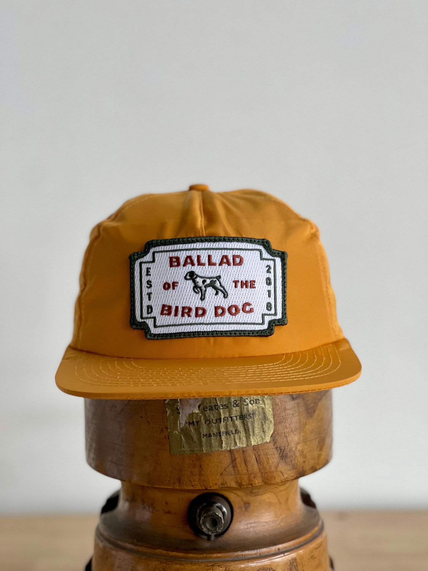 The Ballad of the Bird Dog Shop Hat | Neon Revival is a yellow cap featuring a white patch with red "Ballad of the Bird Dog" text, displayed on a wooden stand to evoke the vibrant spirit of downtown Brenham.