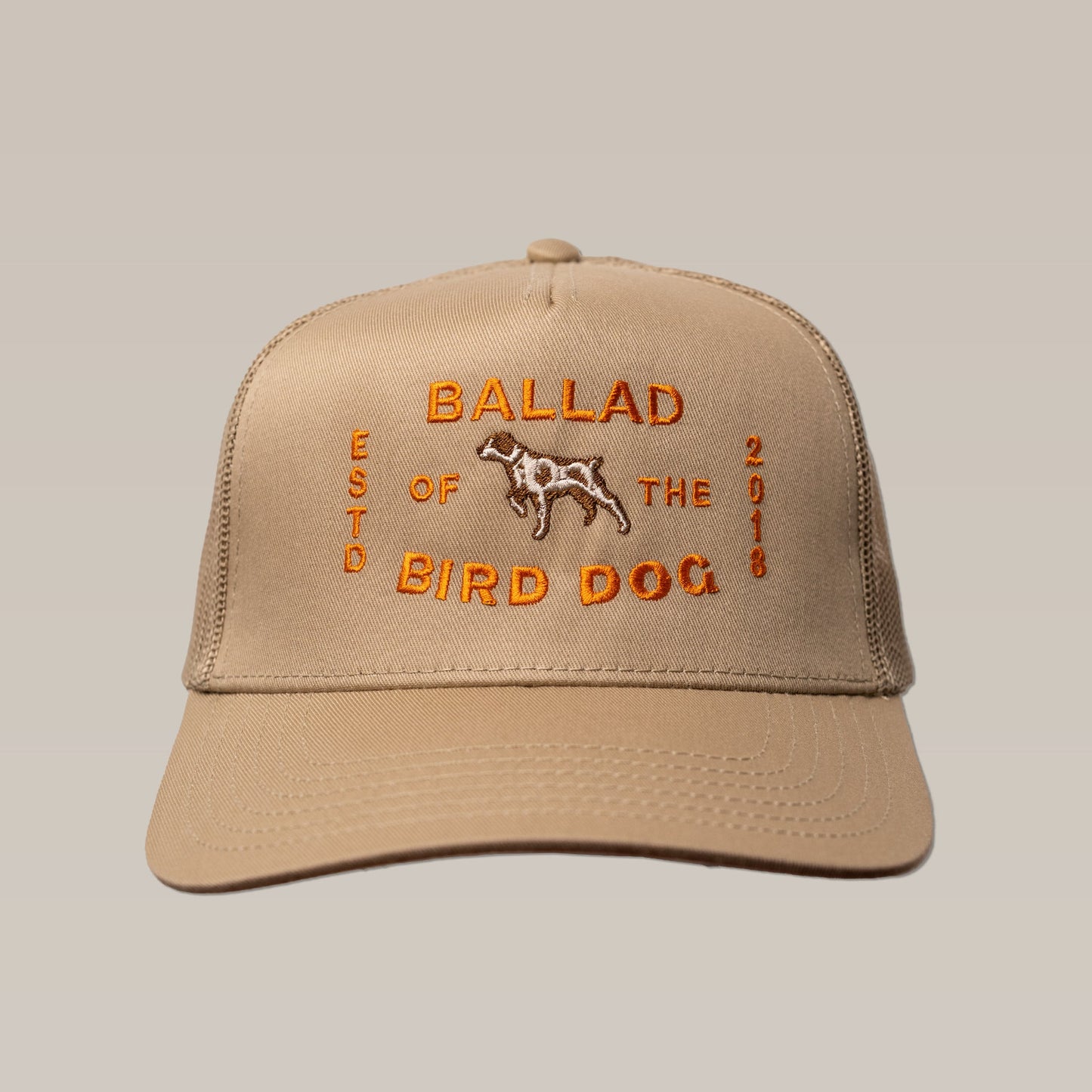 The "Shop Hat | Neon Sign" by Ballad of the Bird Dog is a tan trucker hat featuring orange embroidery with the brand name, a dog illustration, and "ESTD 2018" on the front.