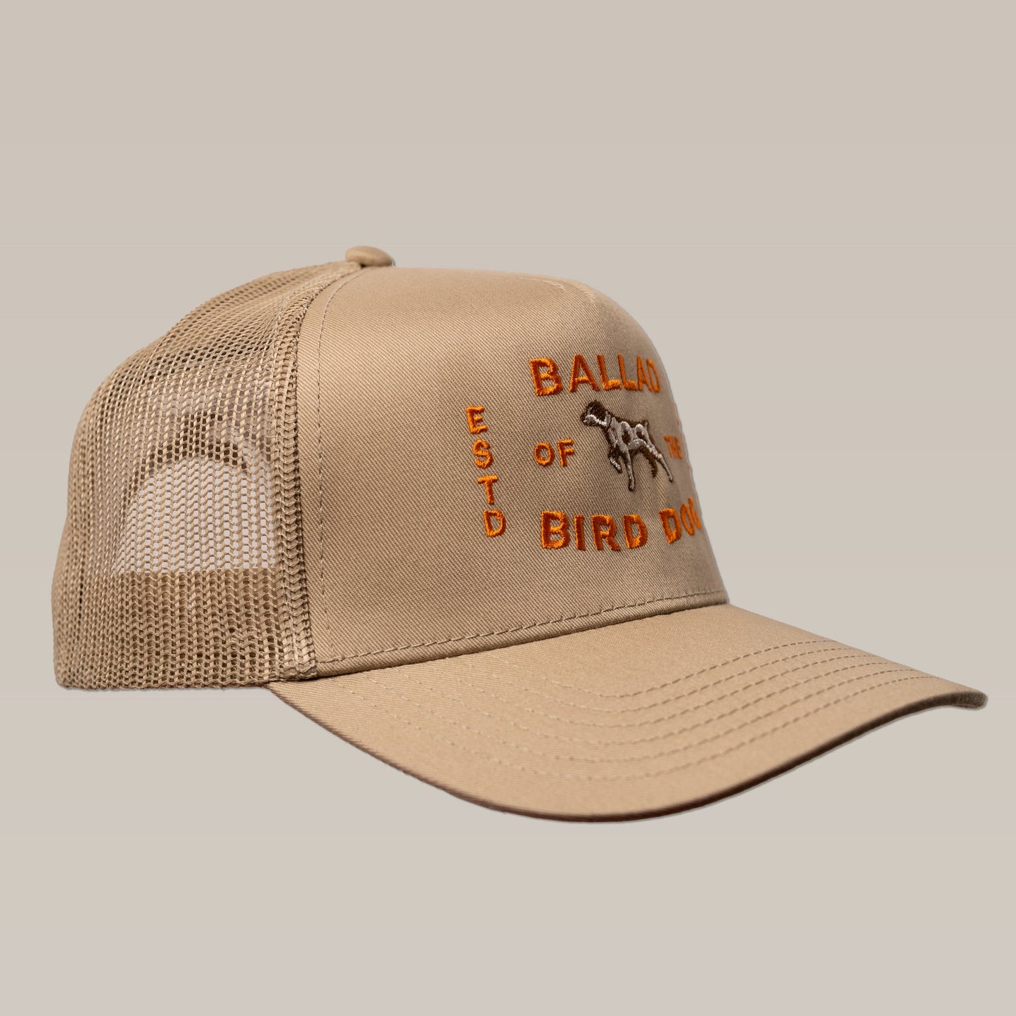 The Ballad of the Bird Dog Shop Hat | Neon Sign is a tan mesh baseball cap featuring orange embroidered text and a dog graphic on the front panel, shown from the left side.