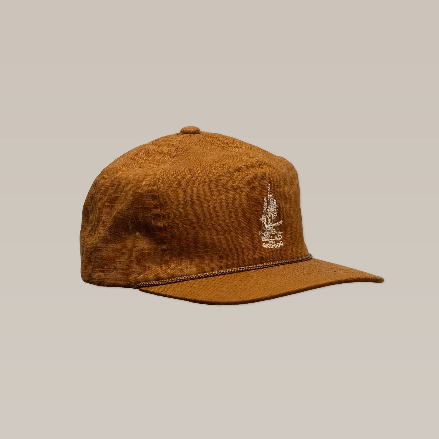 The Ballad of The Bird Dog Shop Hat | Roadrunner Saguaro is a brown fabric baseball cap with Southwest-inspired style, featuring a white embroidered front design and decorative cord above the visor, shown on a plain light background.
