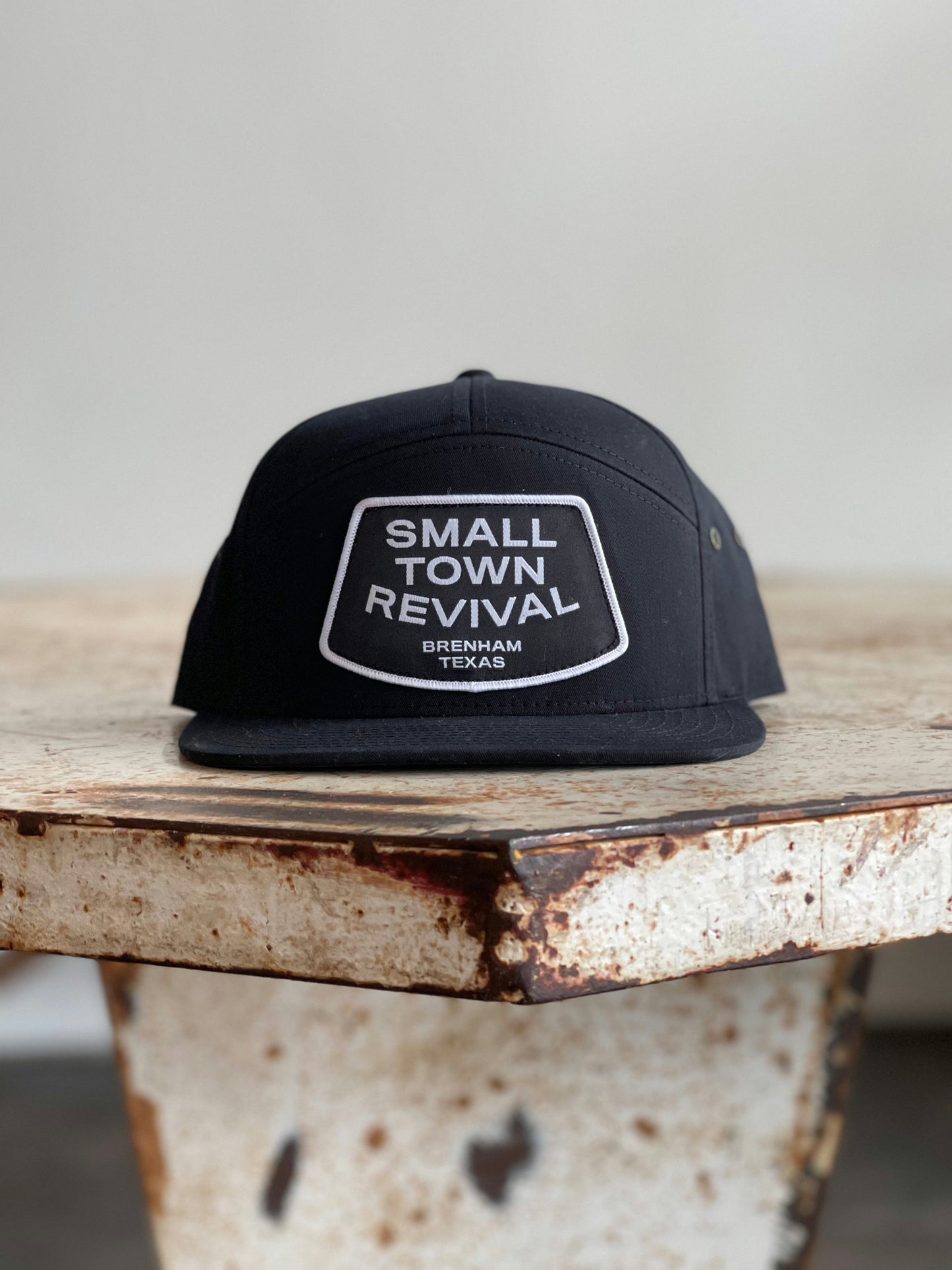 The Shop Hat | Small Town Revival Patch by Ballad of the Bird Dog—a black cap with a white "Small Town Revival Brenham Texas" patch—rests on a rustic table, ideal for Brenham pride and small town hat fans.