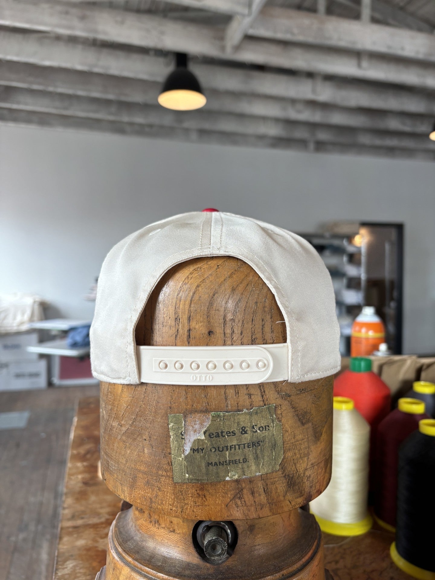 The Shop Hat | Texas Spirit by Ballad of the Bird Dog is a white adjustable cap displayed on a wooden hat form in a workshop, surrounded by sewing supplies and spools of thread.