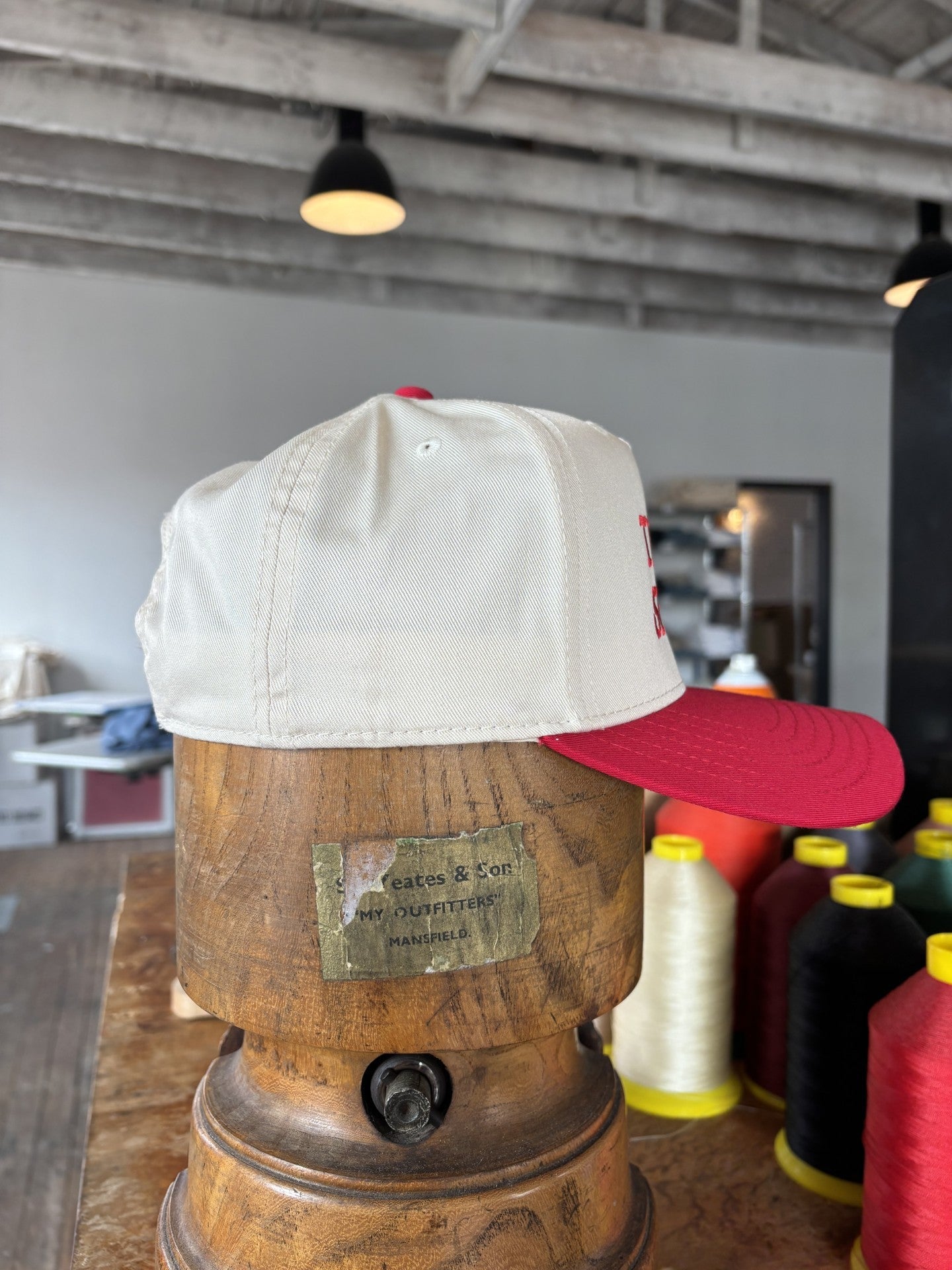 The Shop Hat | Texas Spirit by Ballad of the Bird Dog is a red and white baseball cap shown on a wooden stand, surrounded by colorful spools of thread in a workshop setting.