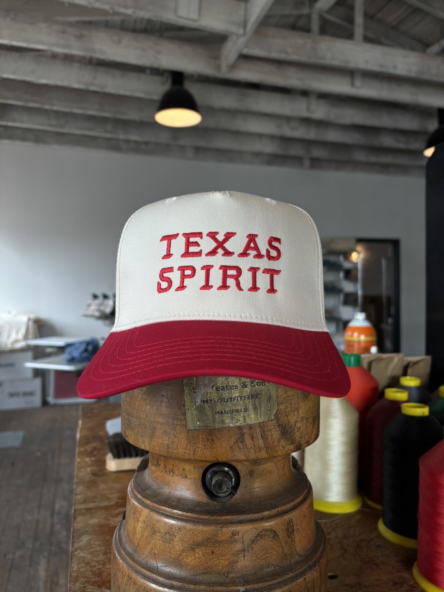The Ballad of the Bird Dog Shop Hat | Texas Spirit features "TEXAS SPIRIT" embroidered in red on a white and red trucker cap, displayed on a wooden stand with sewing thread spools and a workshop in the background.
