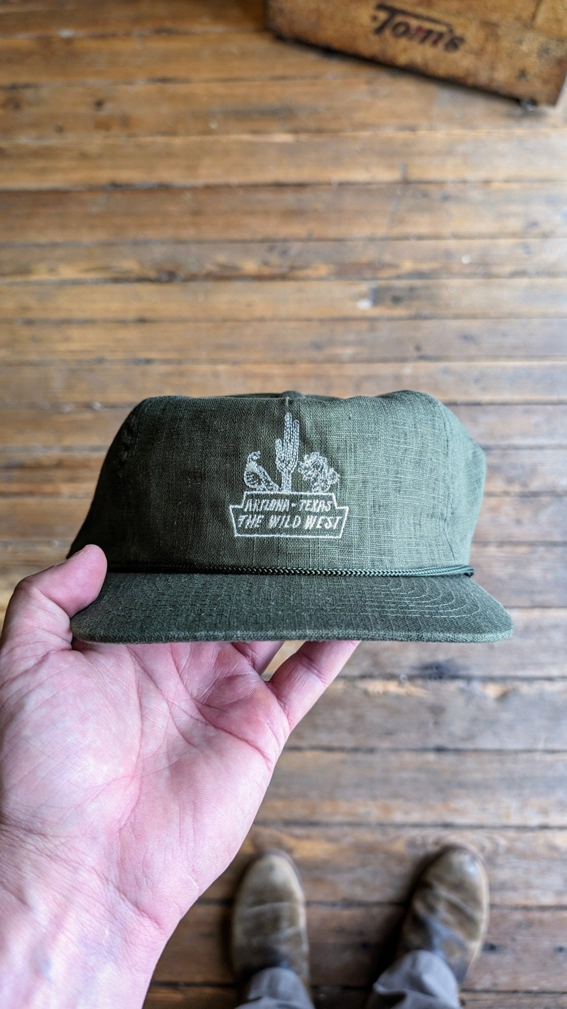 A hand holds the Shop Hat | The Wild West by Ballad of the Bird Dog in olive green, with a white embroidered cactus and "Arizona-Texas The Wild West," part of the Desert Series Collection, shown over wooden floorboards.