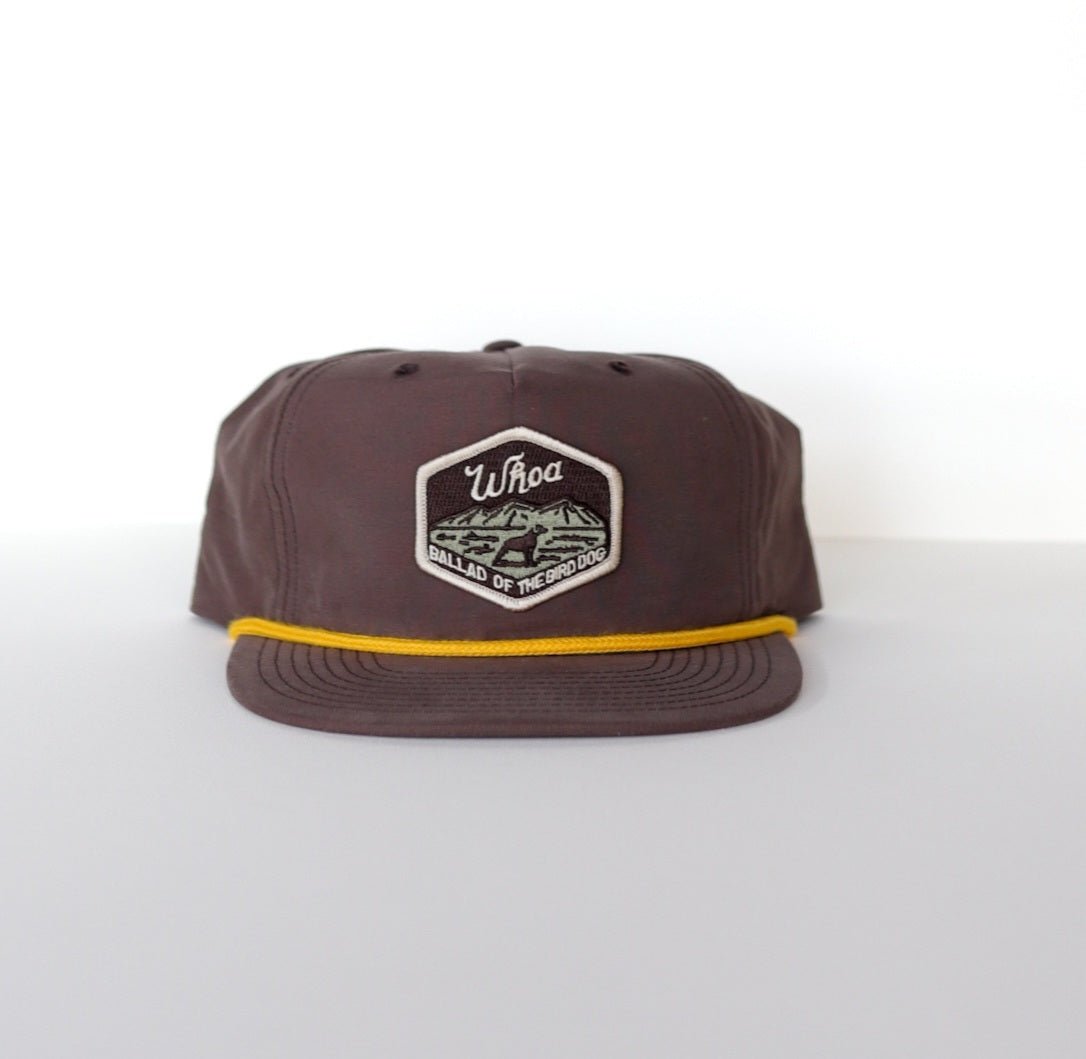 The Shop Hat | Whoa by Ballad of the Bird Dog features a brown snapback with yellow trim and a hexagonal "Whoa Ballad of Incognito" patch—ideal for bird hunting or adventures with your loyal bird dog.