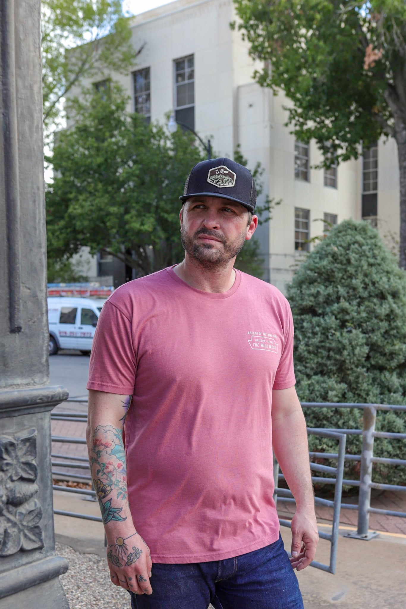 A tattooed man in a pink t-shirt and the Shop Hat | Whoa by Ballad of the Bird Dog stands outside near trees and a building, looking to the side.