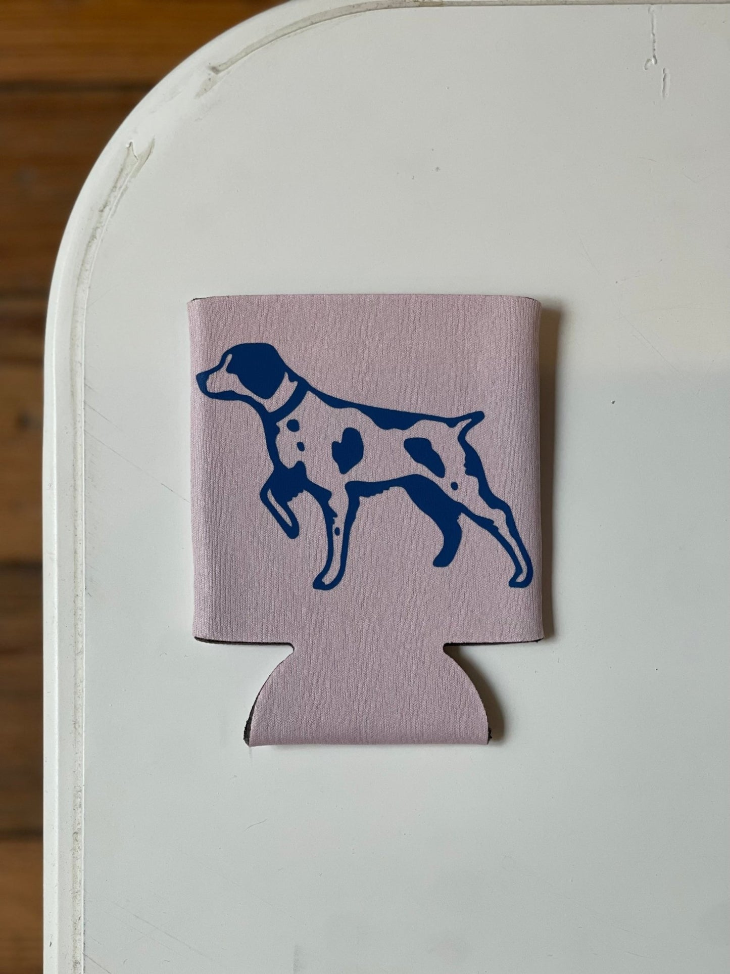The Ballad of the Bird Dog Shop Koozie, featuring a blue dog silhouette, rests on a white tabletop.