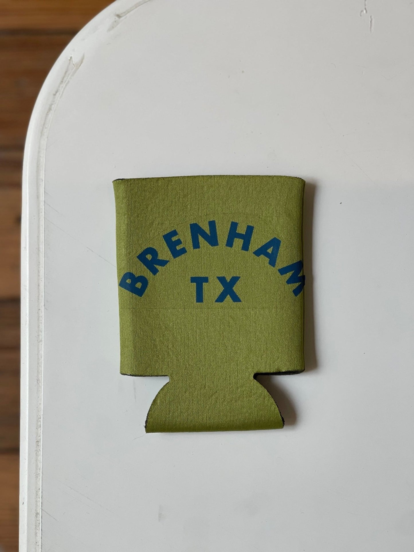 The Shop Koozie by Ballad of the Bird Dog is a green drink koozie featuring "BRENHAM TX" in blue text, shown on a white surface.