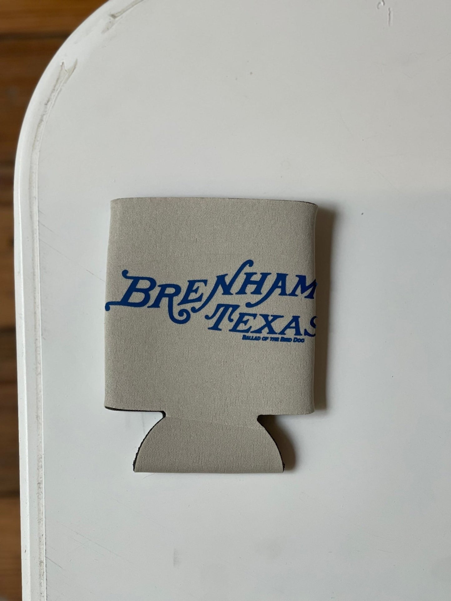 A gray Ballad of the Bird Dog Shop Koozie featuring "Brenham Texas" in blue text rests on a white surface.