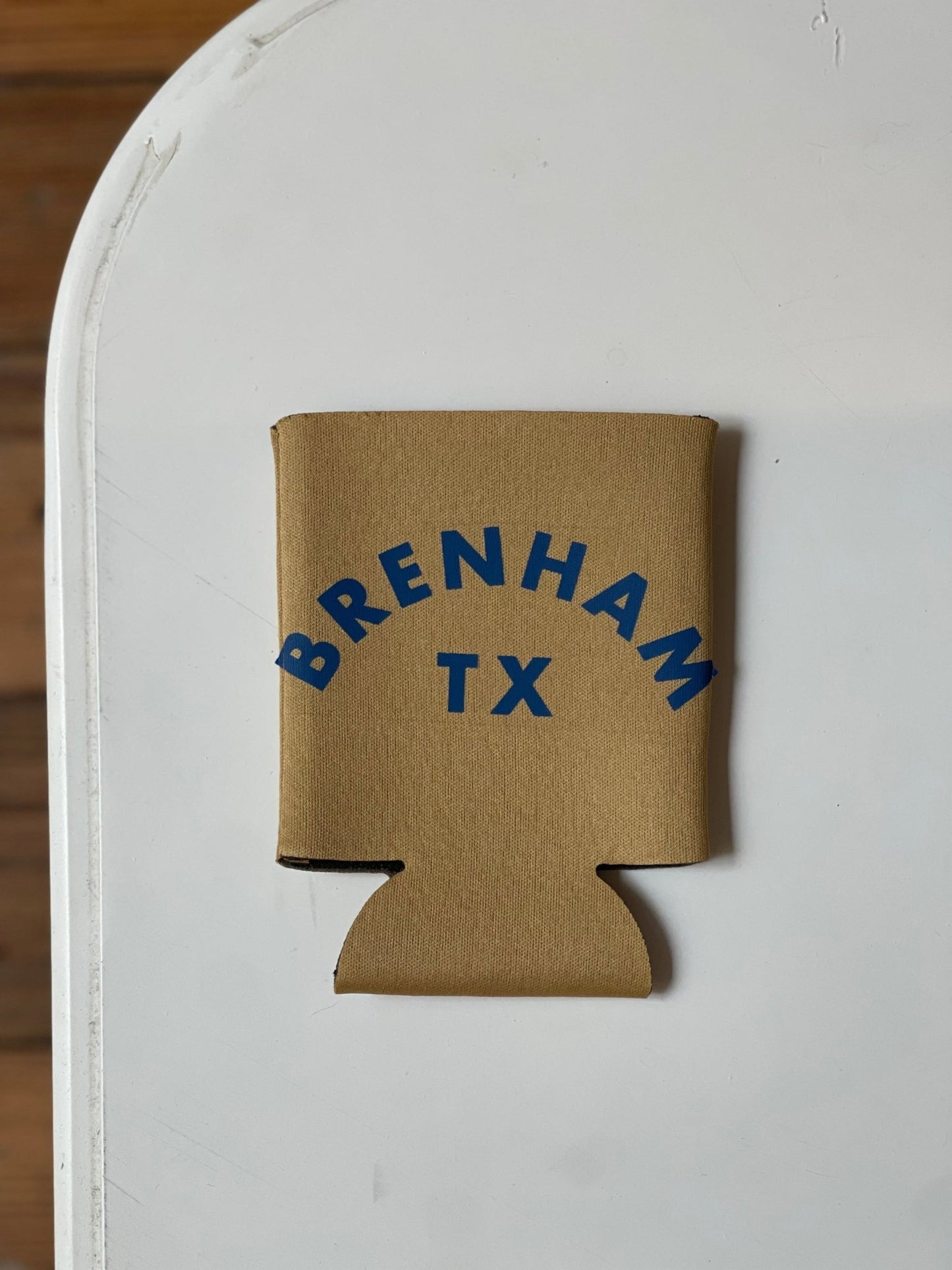 A Ballad of the Bird Dog Shop Koozie featuring "BRENHAM TX" in blue letters rests on a white surface.