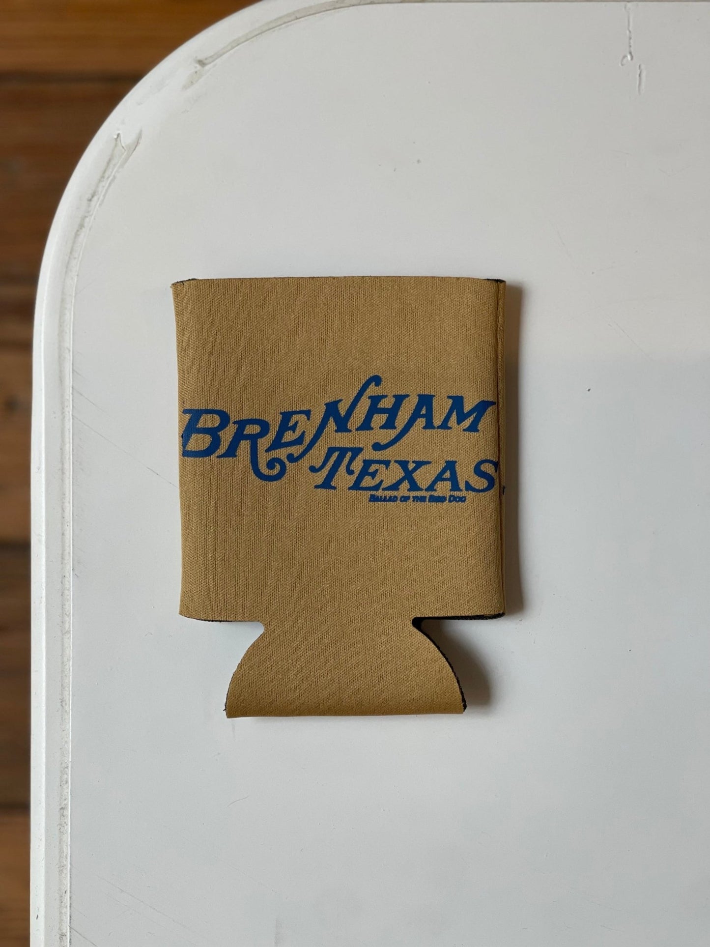 The Shop Koozie by Ballad of the Bird Dog is a tan drink holder with blue "Brenham Texas" text, displayed on a white surface.