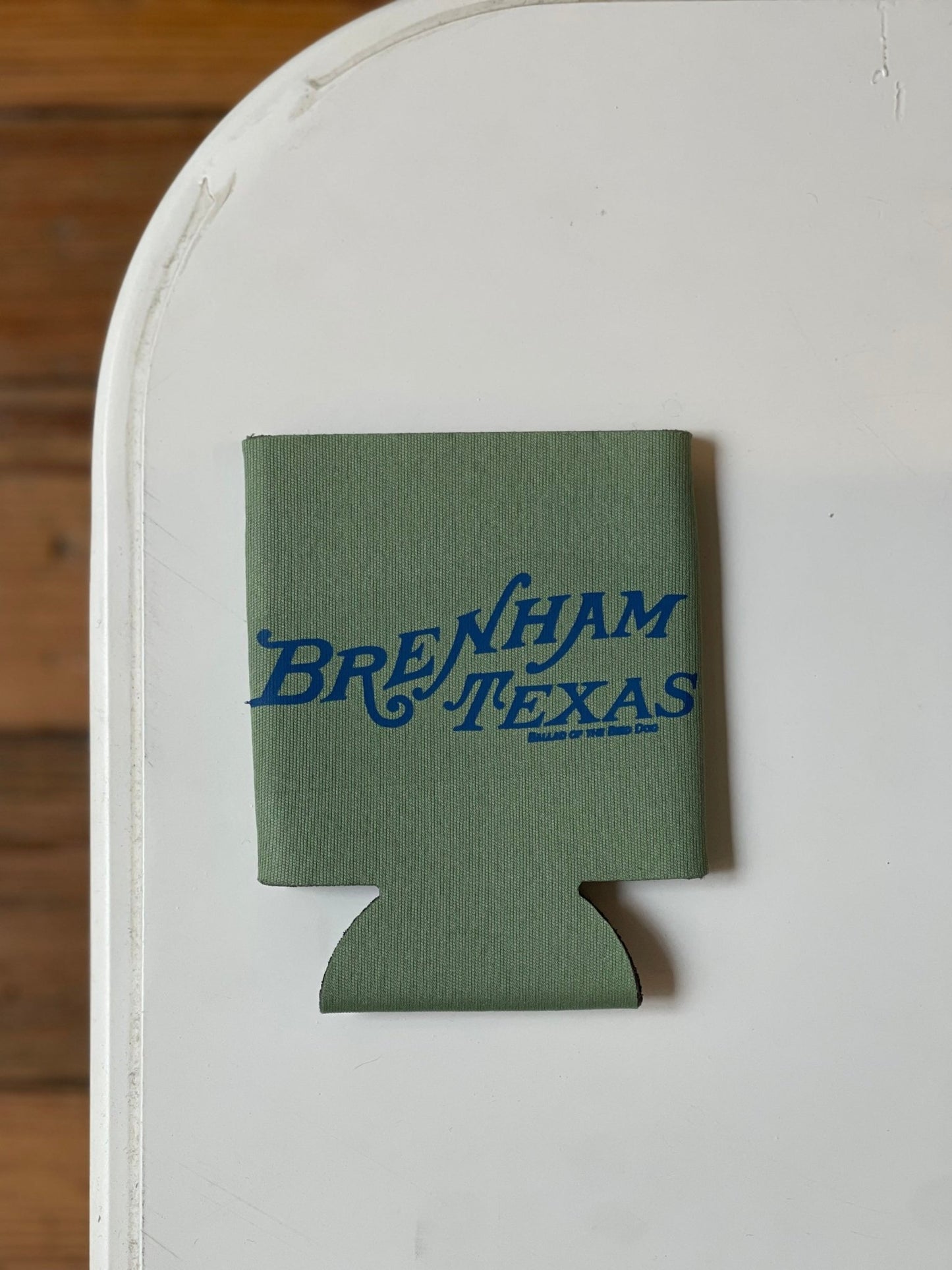 A Shop Koozie by Ballad of the Bird Dog, featuring green material and blue "Brenham Texas" text, sits on a white surface with a wooden floor in the background.