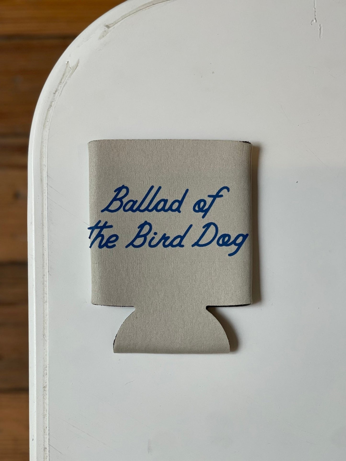 The Ballad of the Bird Dog Shop Koozie, featuring blue text, is displayed on a white surface.
