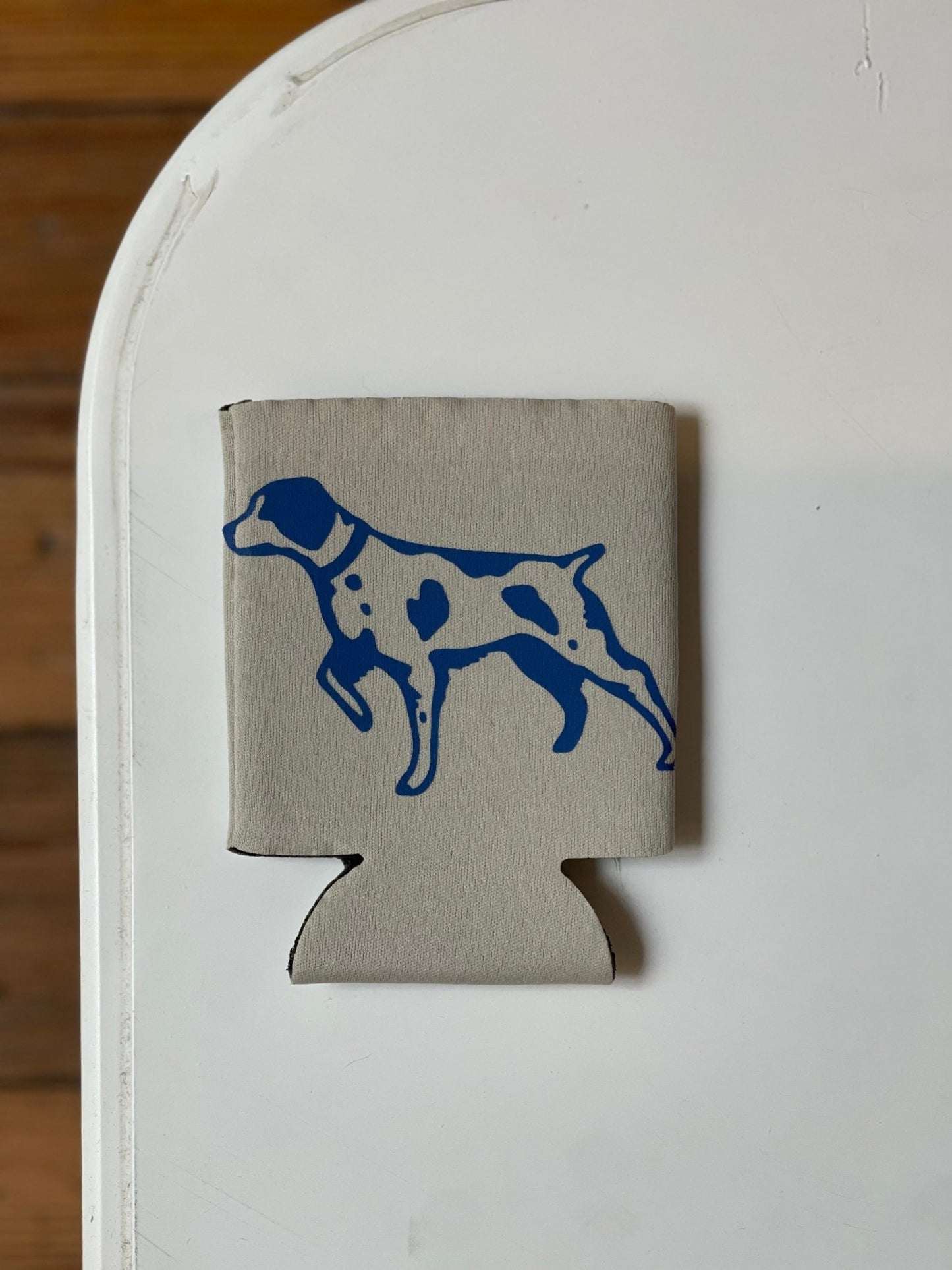 The Shop Koozie by Ballad of the Bird Dog—a gray can cooler with a blue dog silhouette—rests on a white tabletop, with part of a wood floor visible in the background.
