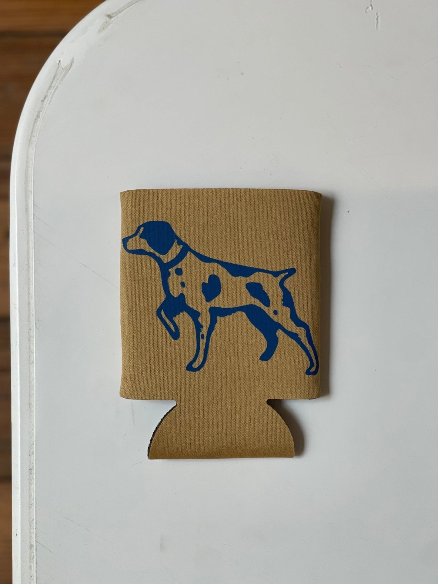 A brown "Shop Koozie | Ballad of the Bird Dog" by Ballad of the Bird Dog, featuring a blue dog silhouette, is displayed on a white surface.