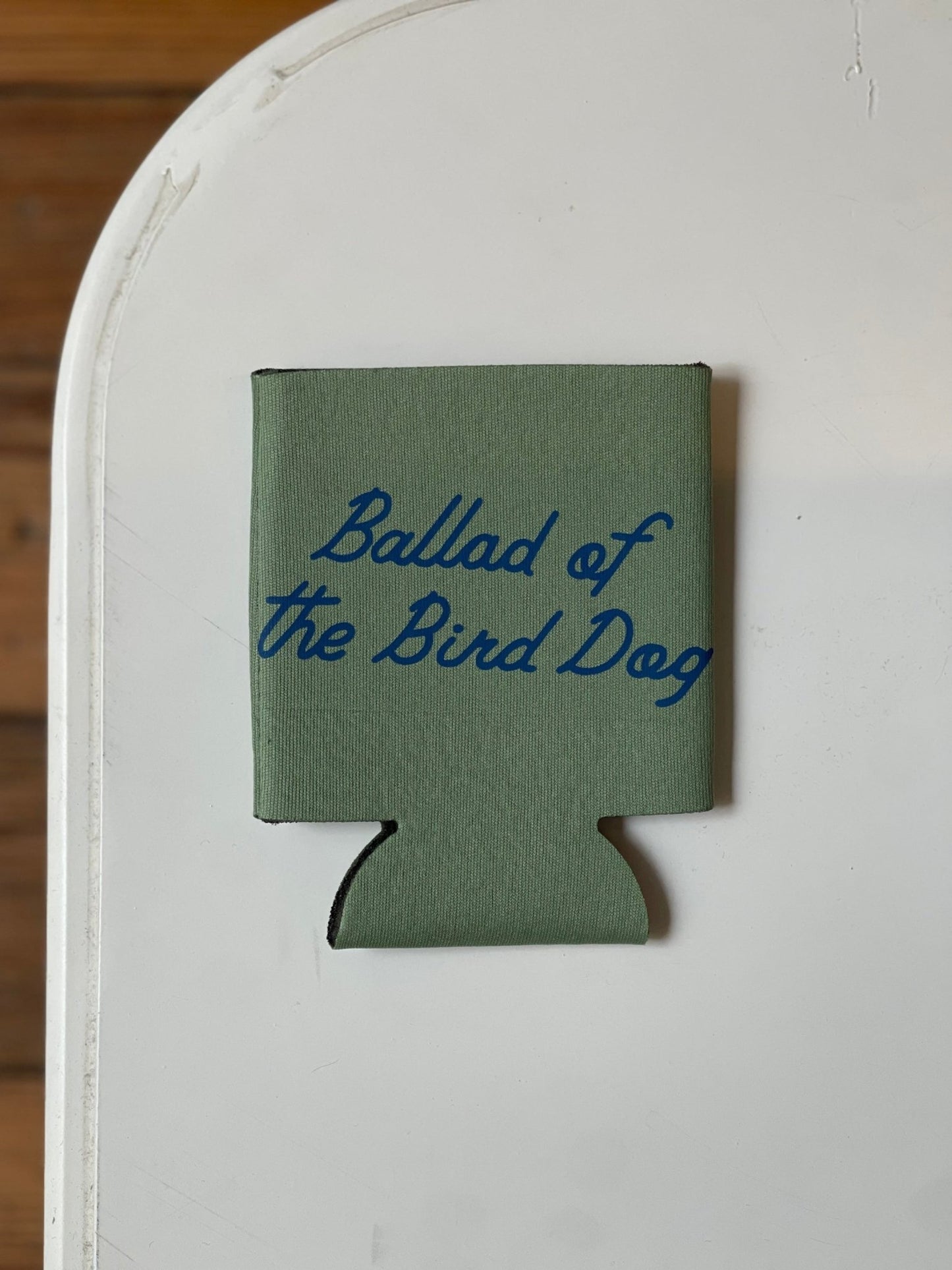 The Shop Koozie | Ballad of the Bird Dog by Ballad of the Bird Dog is a green can cooler featuring "Ballad of the Bird Dog" in blue script, displayed on a white surface.