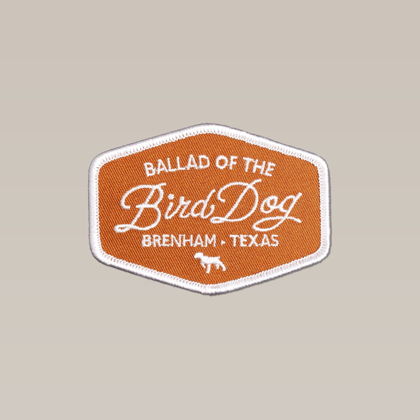 The Bird Dog Badge Patch – Handcrafted Outdoor Emblem by Ballad of the Bird Dog is an orange hexagonal patch with white trim, featuring “Ballad of the Bird Dog, Brenham, Texas” and a small white dog at the bottom—perfect for your next adventure.