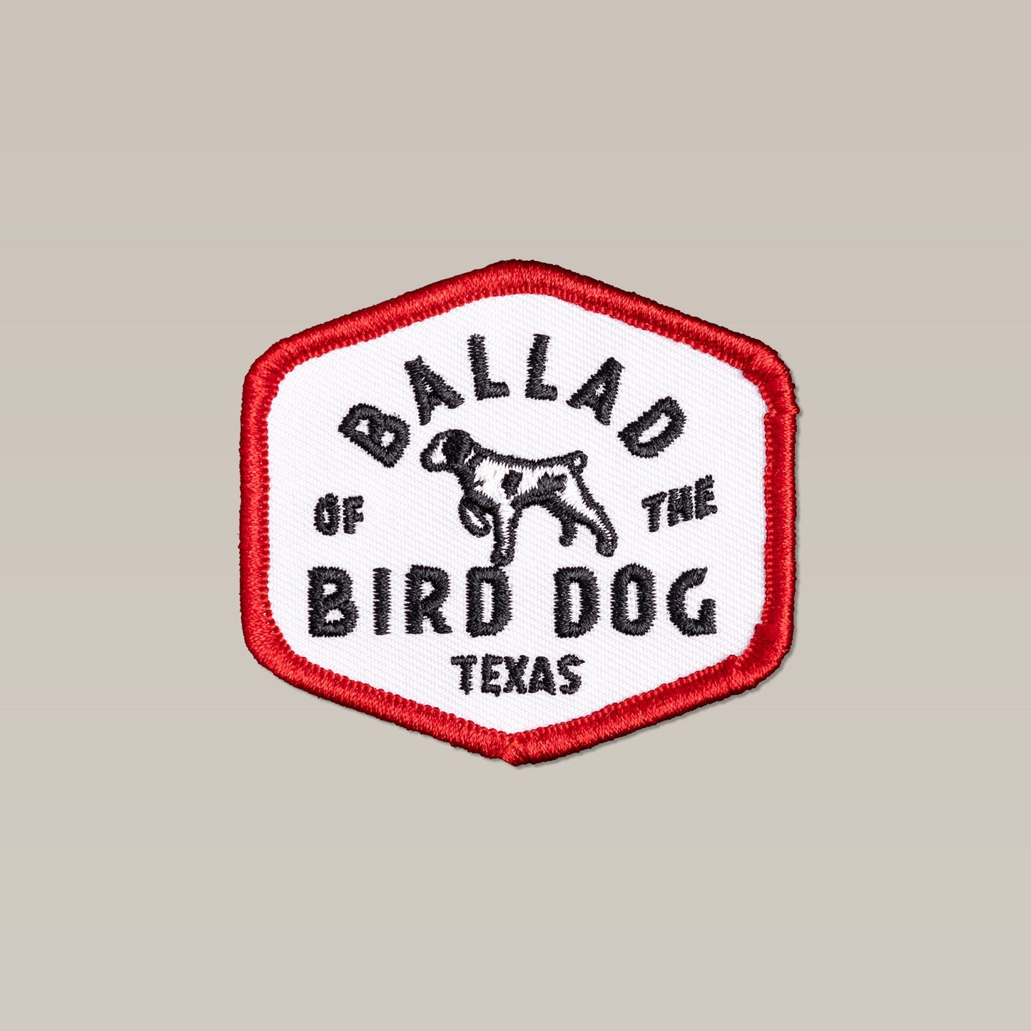 The Shop Patch | Field Companion by Ballad of the Bird Dog is a hexagonal embroidered patch with a red border, dog graphic, and "Ballad of the Bird Dog Texas" in black letters—a must-have accessory for fans.