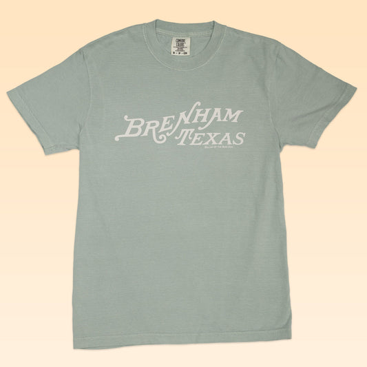 The Shop Shirt | Brenham Texas Heritage Script by Ballad of the Bird Dog features "Brenham Texas" in white script across a light blue shirt, shown on a beige background.