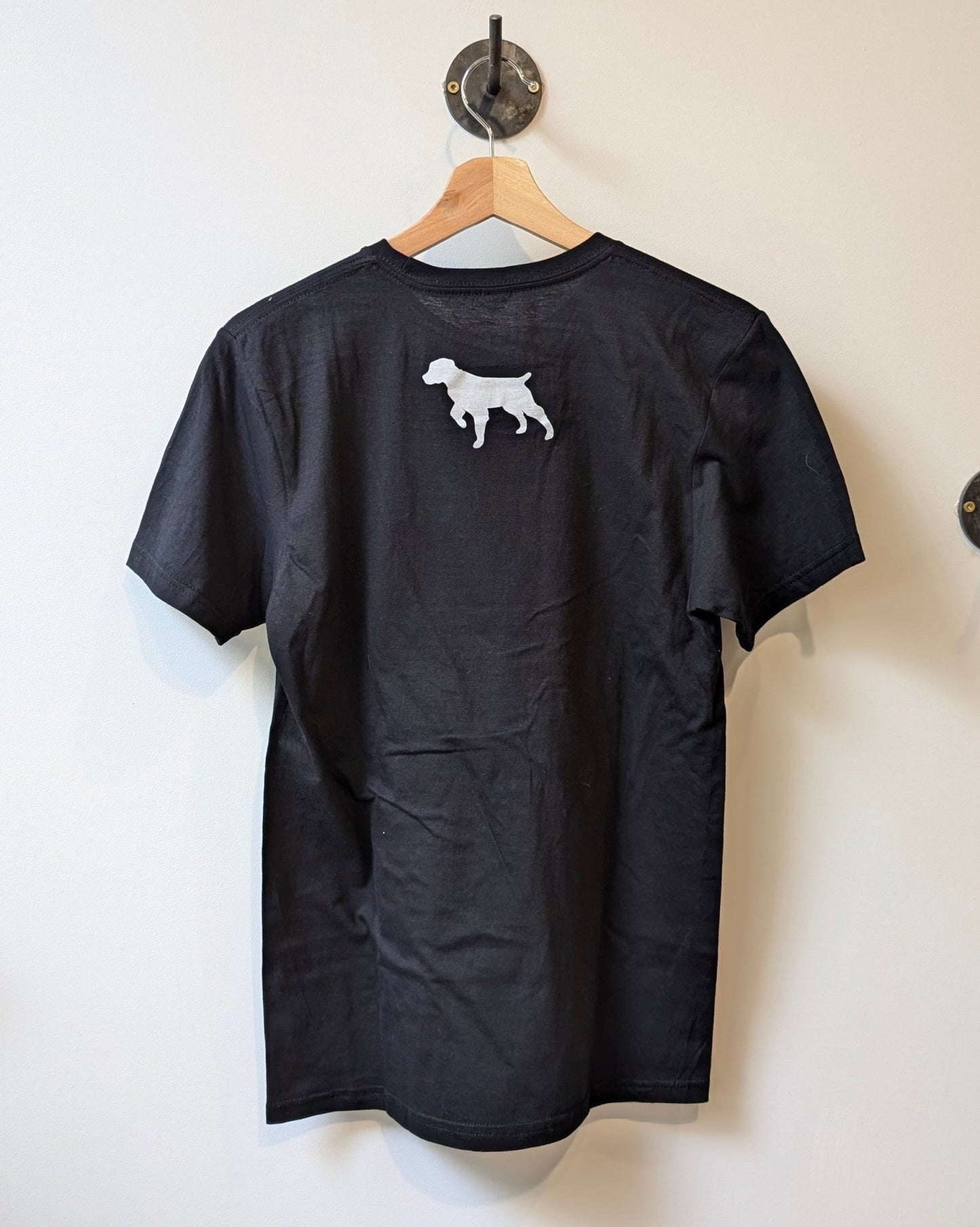 The black Shop Shirt | Brenham Varsity by Ballad of the Bird Dog hangs on a wooden hanger, showcasing a small white dog silhouette printed on the upper back.