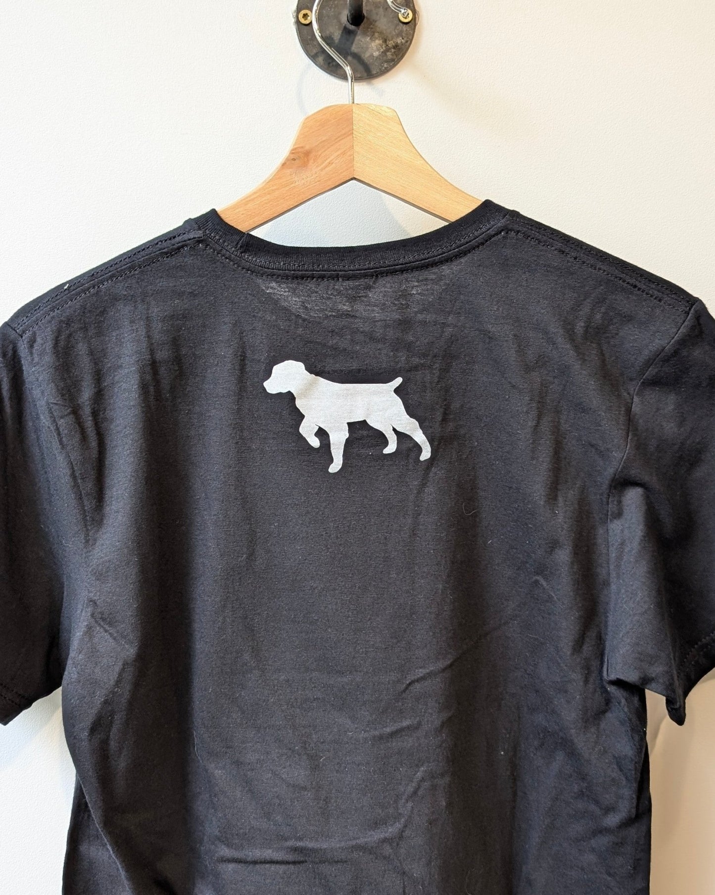 The Ballad of the Bird Dog Shop Shirt | Brenham Varsity is a black collegiate-inspired piece, displayed on a wooden hanger, with a small white dog silhouette featured on the upper back.