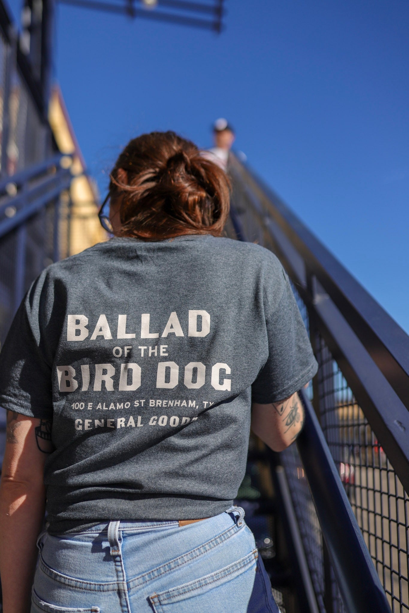 Wearing the Shop Shirt | General Goods by Ballad of the Bird Dog, someone climbs an outdoor metal staircase on a sunny day, channeling the relaxed style of Brenham’s top general goods store.