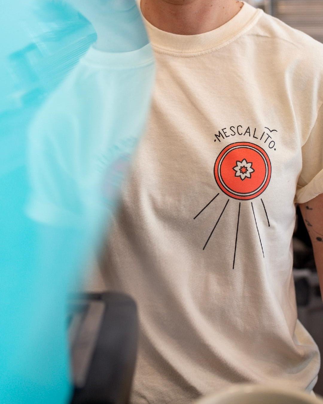 Person wearing the Shop Shirt | Hawks by Mescalito Coffee, featuring a red circular "Mescalito" graphic on a cream background, inspired by West Texas. A blue object partially obscures the left side of the image.