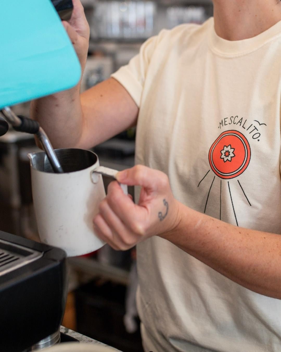 Wearing the cream Shop Shirt | Hawks by Mescalito Coffee, featuring a bold red circular "MESCALITO" graphic, someone steams milk behind the bar—channeling the laid-back West Texas spirit of Mescalito Coffee.