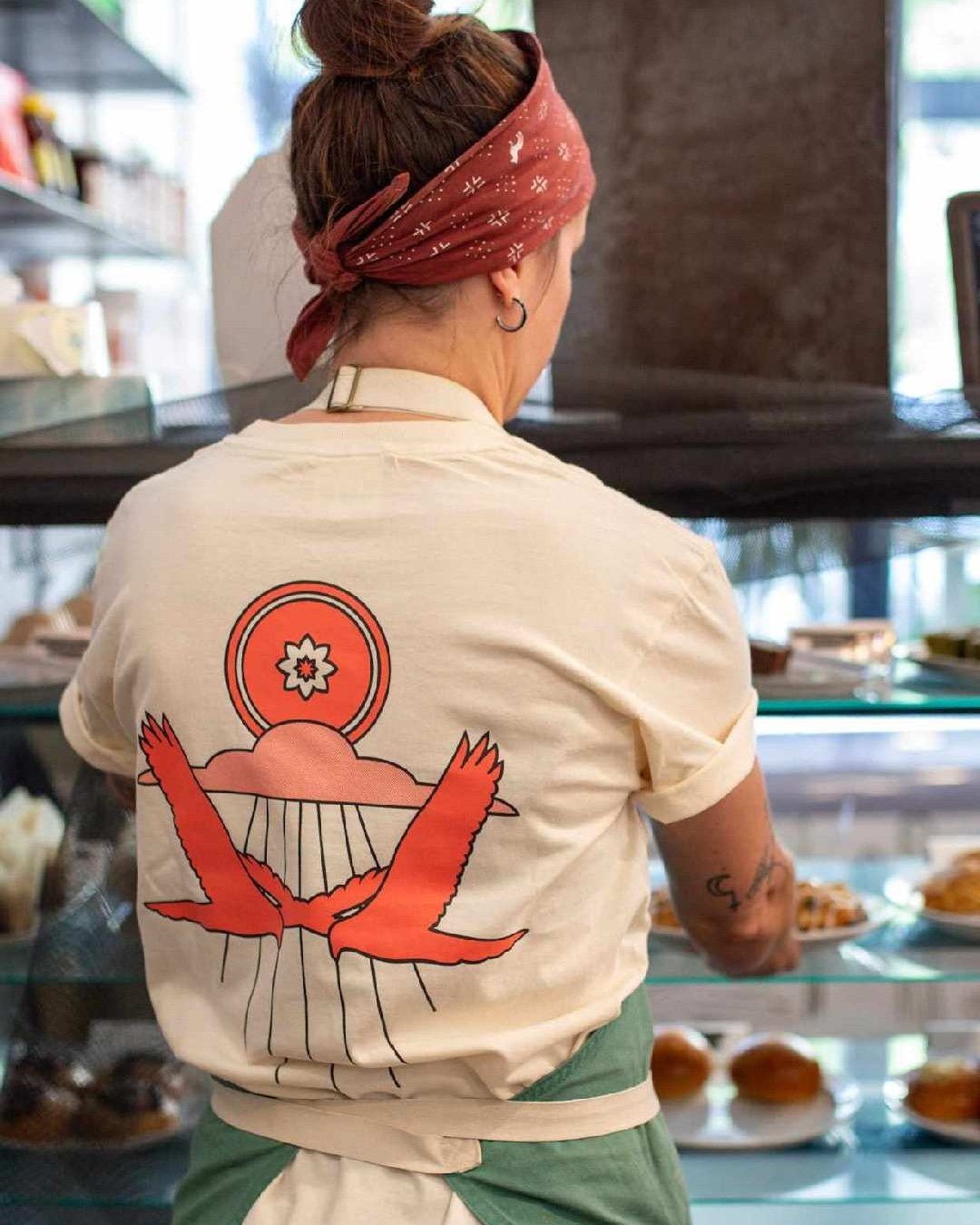 A person in a bandana and apron stands by the pastry case at Mescalito Coffee, sporting the Shop Shirt | Hawks tee with a red bird and sunrise design on the back, perfectly matching the West Texas vibe.