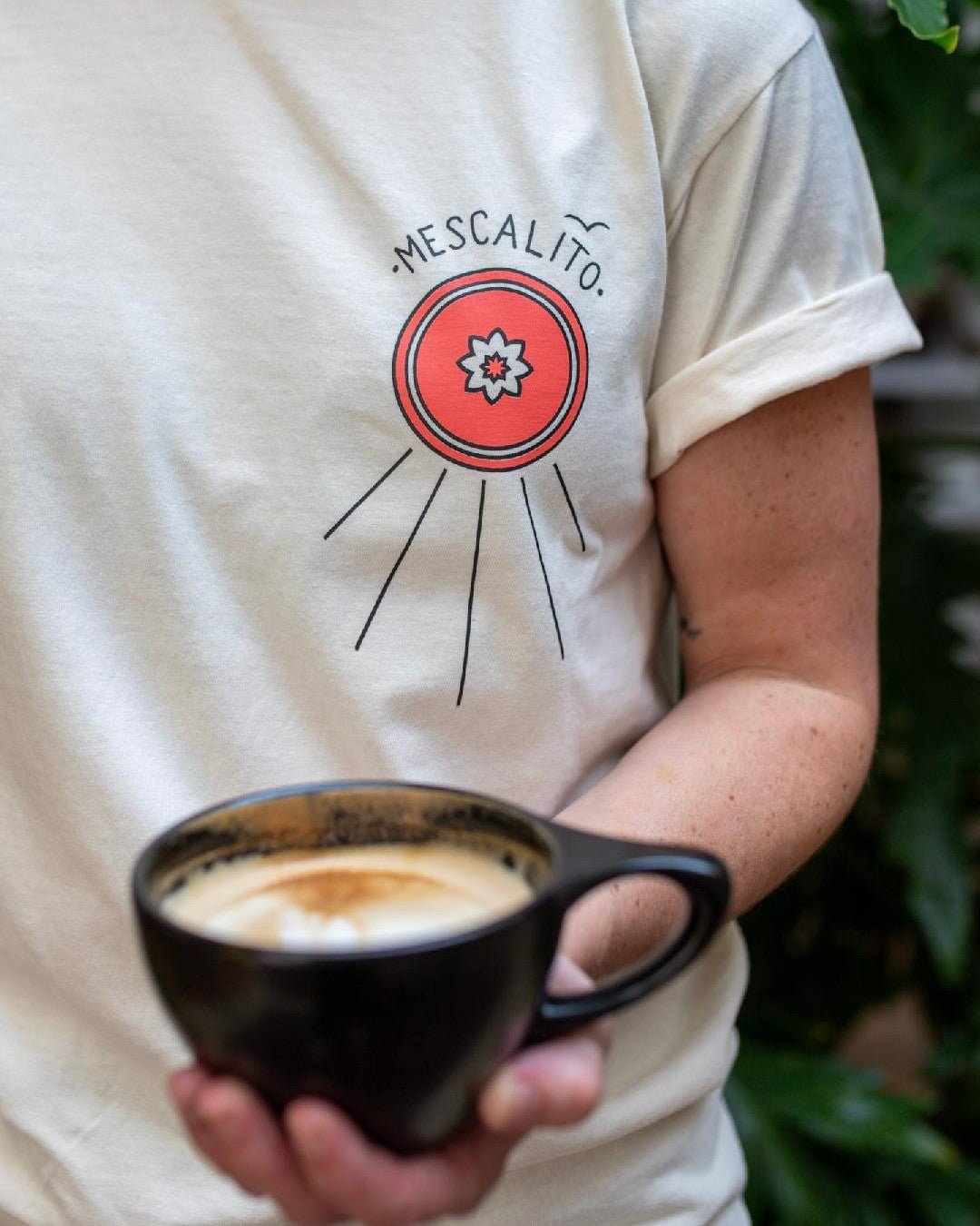 A person in a white "Shop Shirt | Hawks | Mescalito Coffee" tee from Mescalito Coffee holds a black coffee with foam, standing before green plants.