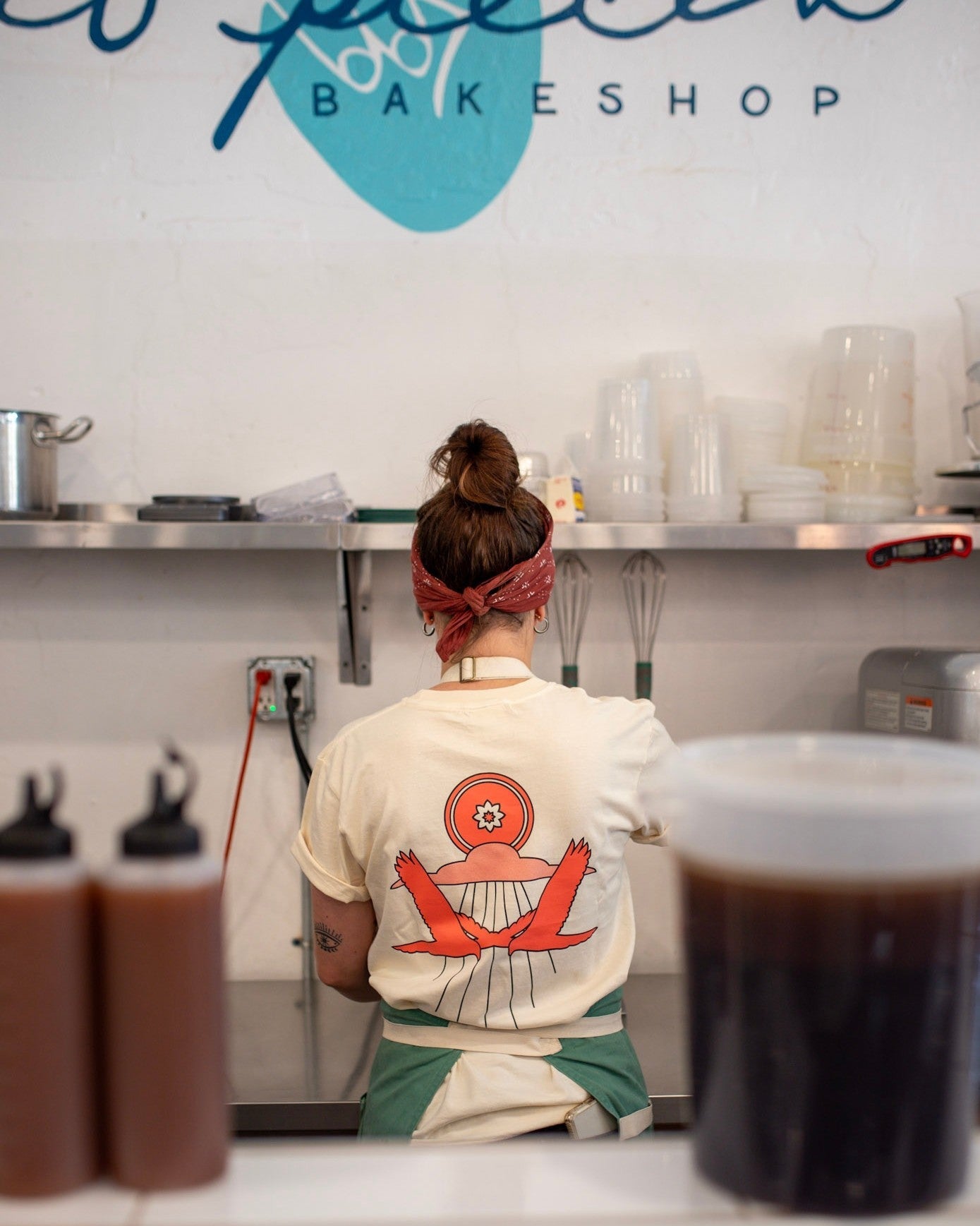 Wearing the Shop Shirt | Hawks by Mescalito Coffee, a person with a bun and bandana stands at a bakeshop counter, exuding a relaxed West Texas vibe as they face away from the camera.