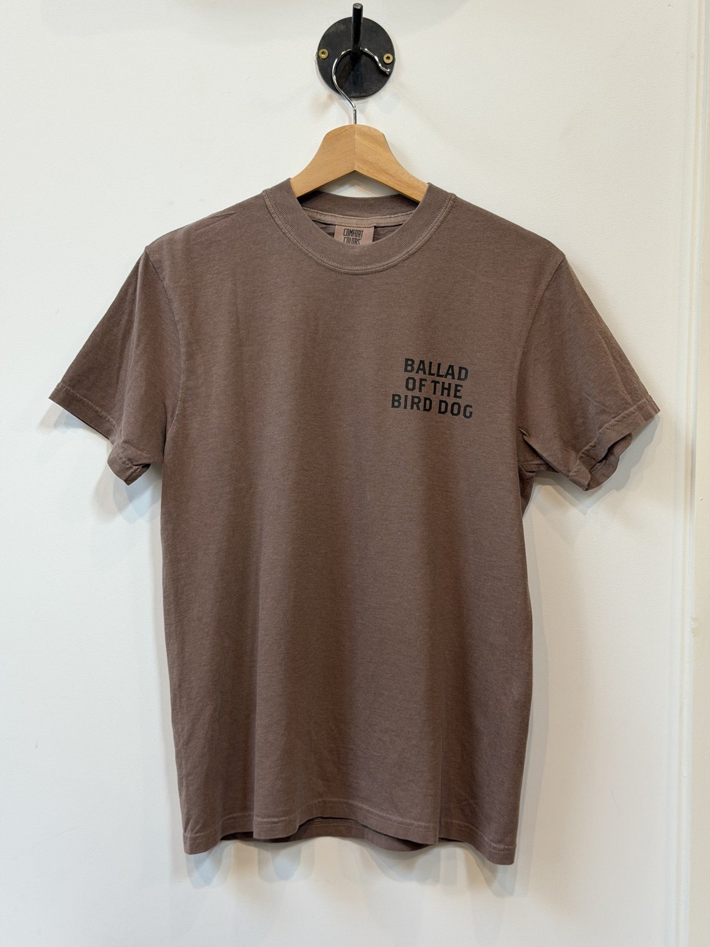 A plain brown Ballad of the Bird Dog "Shop Shirt | Loyal to The Land" t-shirt on a hanger, featuring "BALLAD OF THE BIRD DOG" printed in black on the left chest.