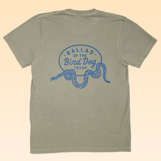 The Shop Shirt by Ballad of the Bird Dog features a light olive green color, blue “BALLAD OF THE BIRD DOG TEXAS” text, and a rattlesnake oval logo graphic on the back.