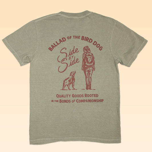 The Shop Shirt | Side By Side by Ballad of the Bird Dog is a green shirt featuring a maroon dog and person graphic with the text "BALLAD OF THE BIRD DOG, Side By Side, QUALITY GOODS ROOTED IN THE BONDS OF COMPANIONSHIP," honoring bird dog bonds.